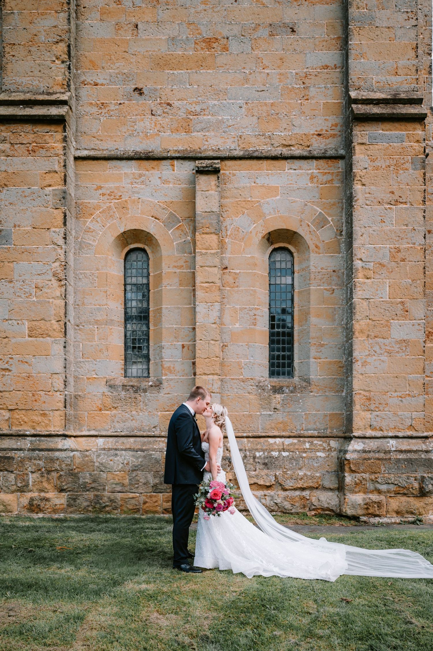 Wedding couple posing against historic stone church wall with arched windows.
