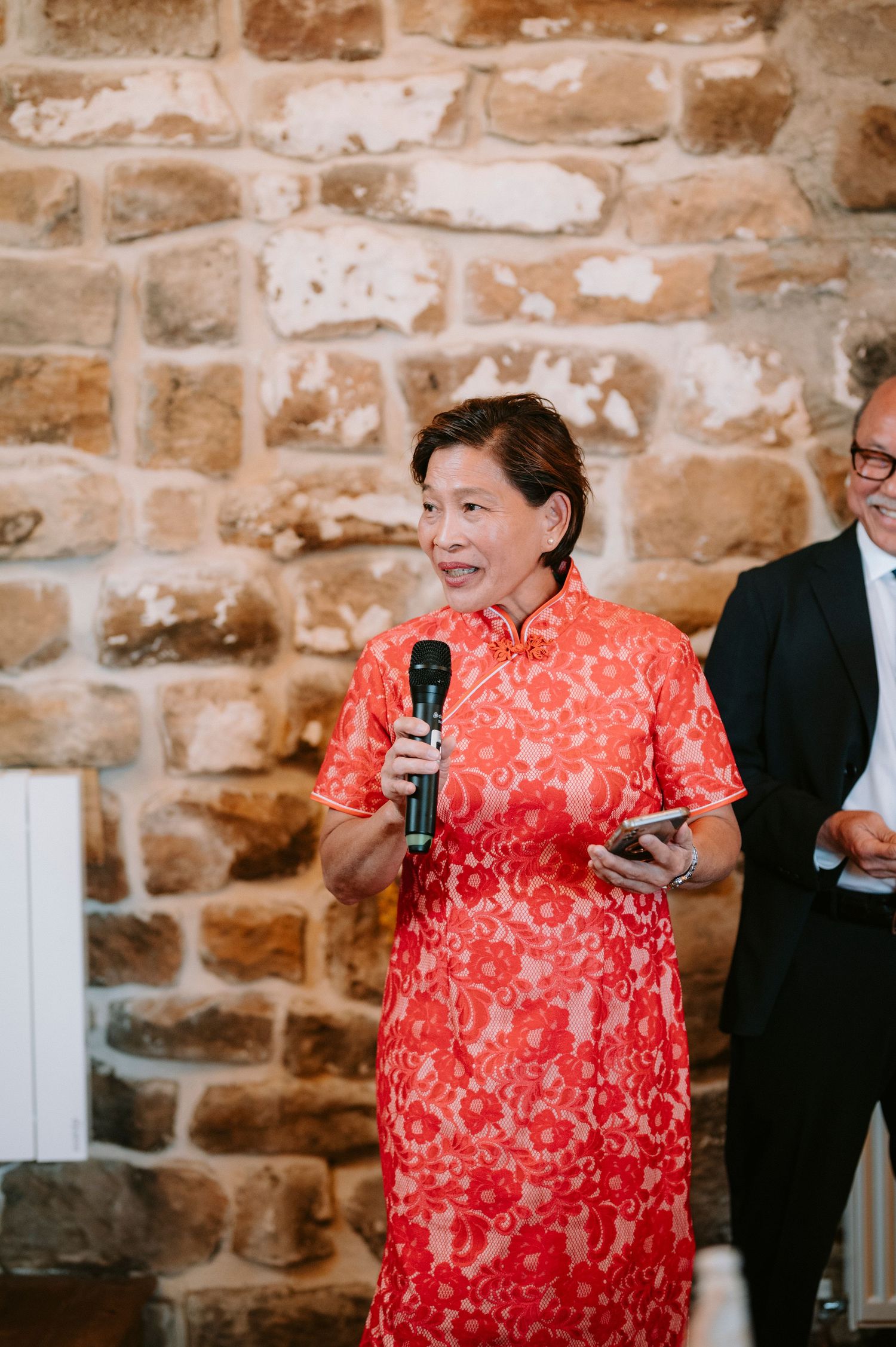 A speaker in a traditional red Chinese qipao dress stands in front of a stone wall during an event.