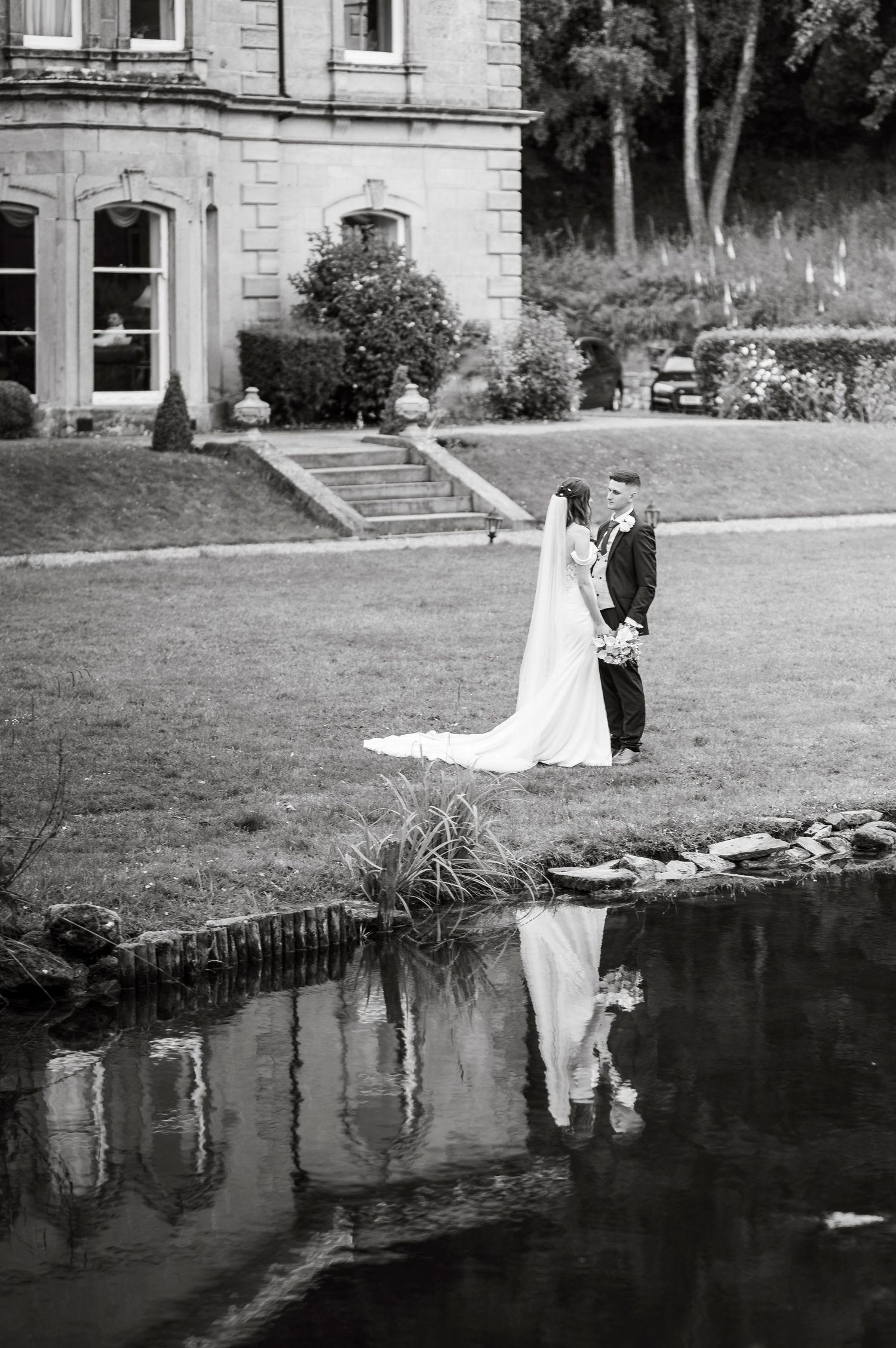 Black and white wedding photography by a reflective pond at an elegant stone estate.