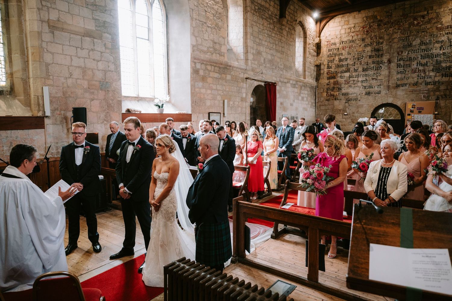 Wedding guests seated in a historic stone church with red chairs and brick walls during a ceremony.