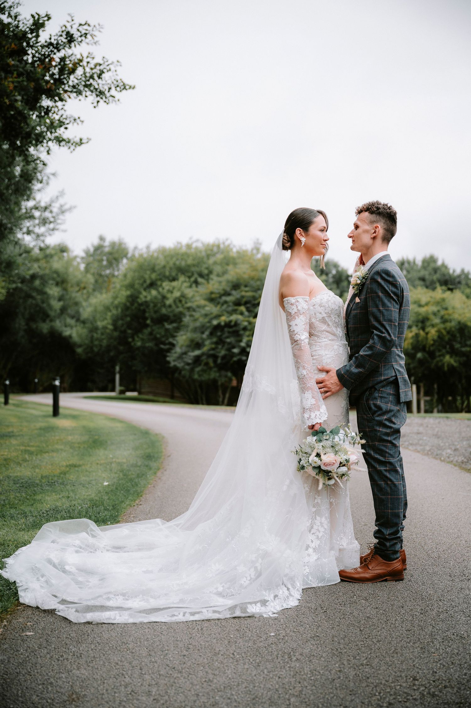 A romantic wedding couple shares intimate moments on a tree-lined path in their elegant wedding attire.