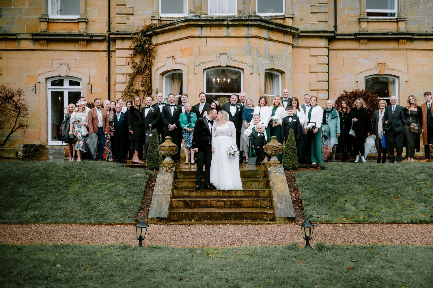 Wedding guests gather on the lawn of a grand stone mansion for a group photo.