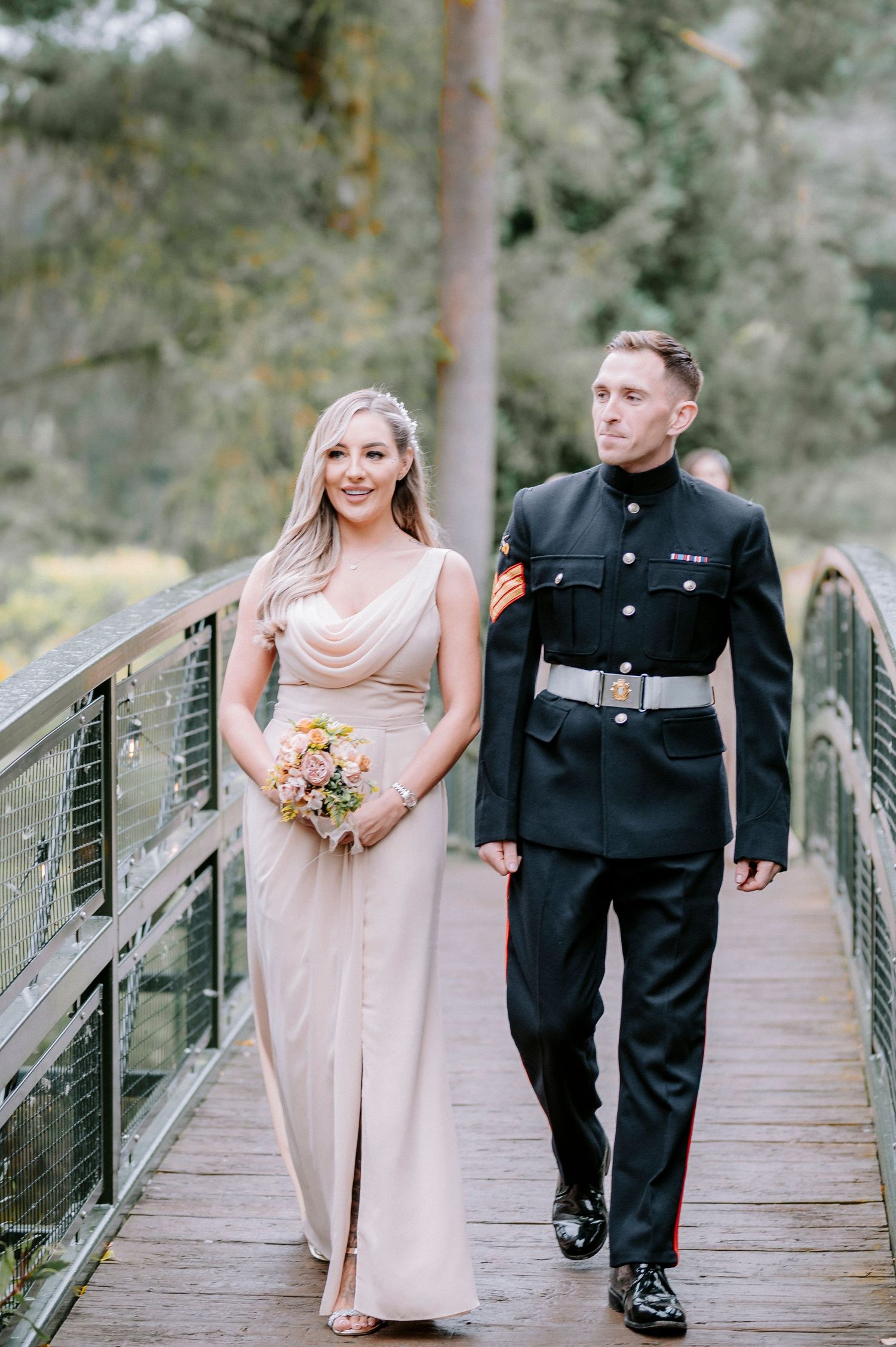 Wedding party members walk across a wooden bridge in formal attire with blush bridesmaid dresses.