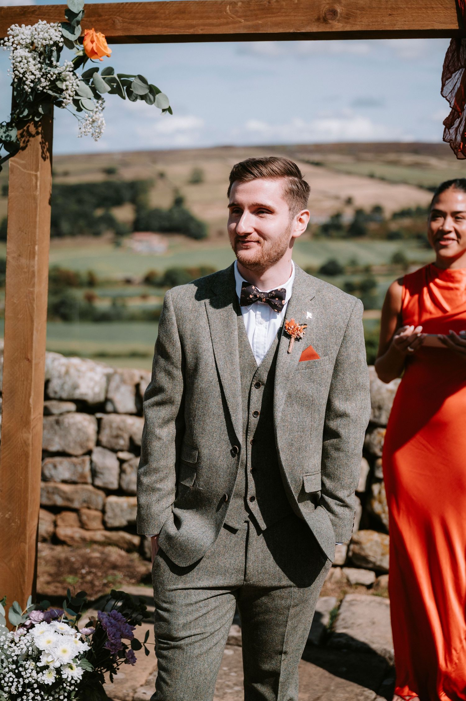 Wedding party members in gray suits and coral dresses stand together against a scenic countryside backdrop.
