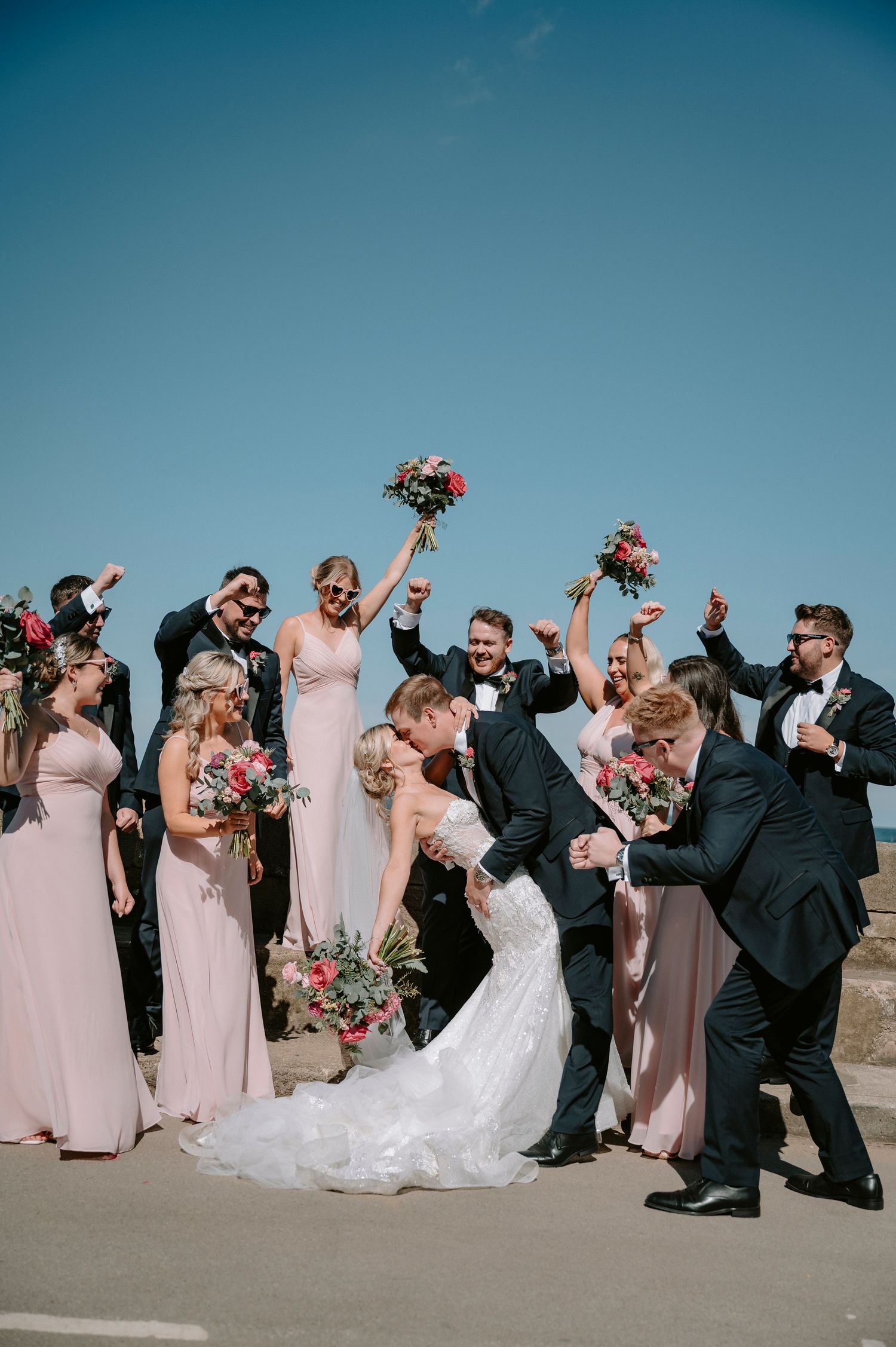 Wedding party cheers with raised bouquets during outdoor celebration on sunny day.