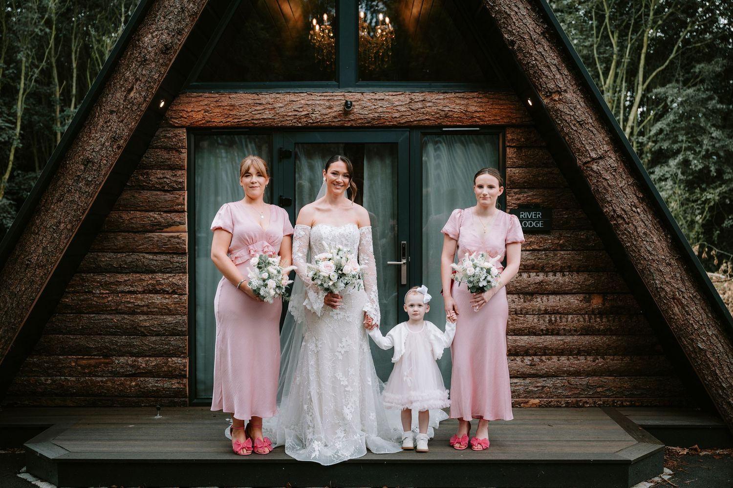 Wedding party in pink dresses poses in front of rustic wooden cabin.