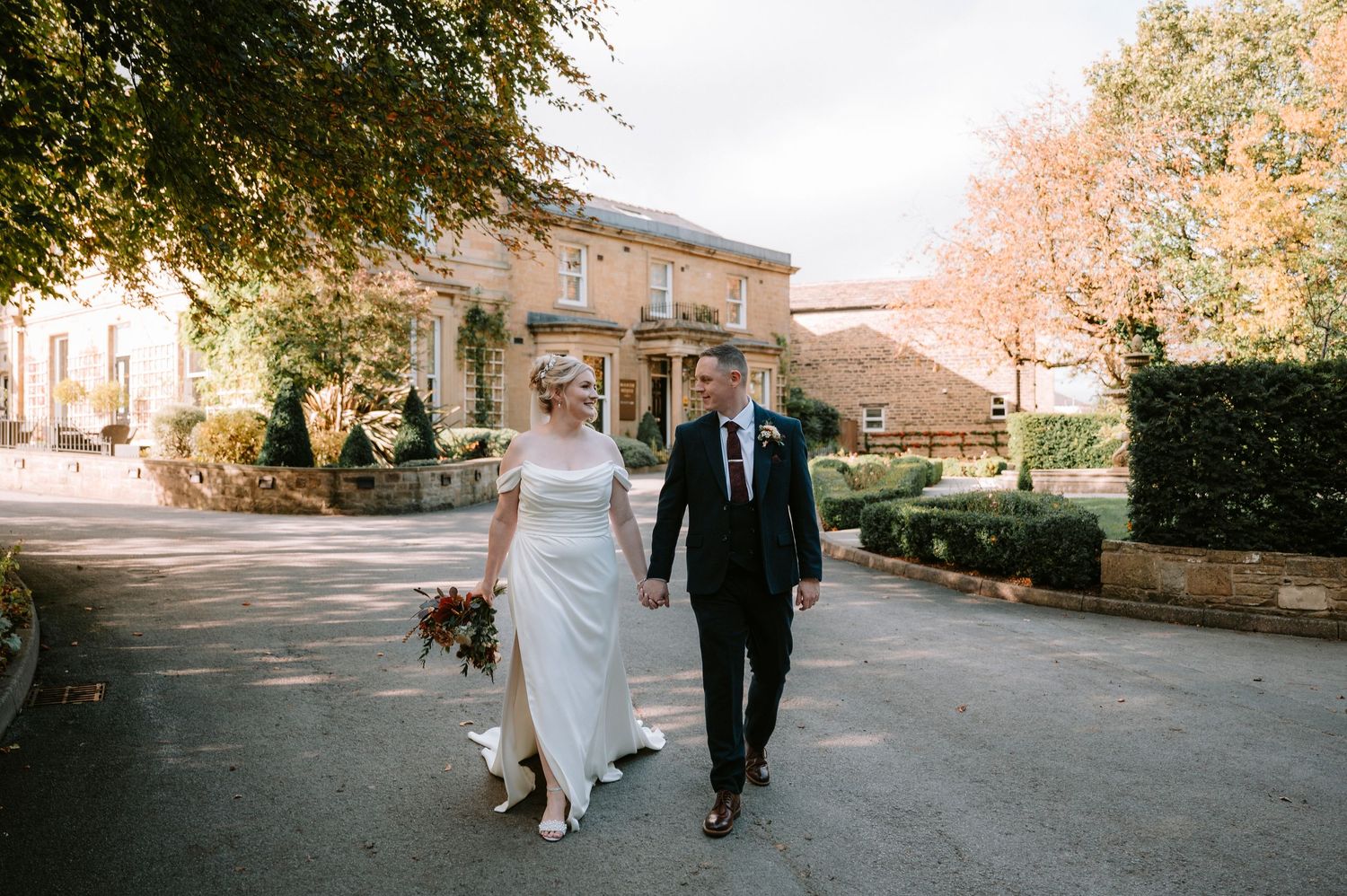 A couple walks together down a tree-lined path in formal attire during an autumn wedding celebration.