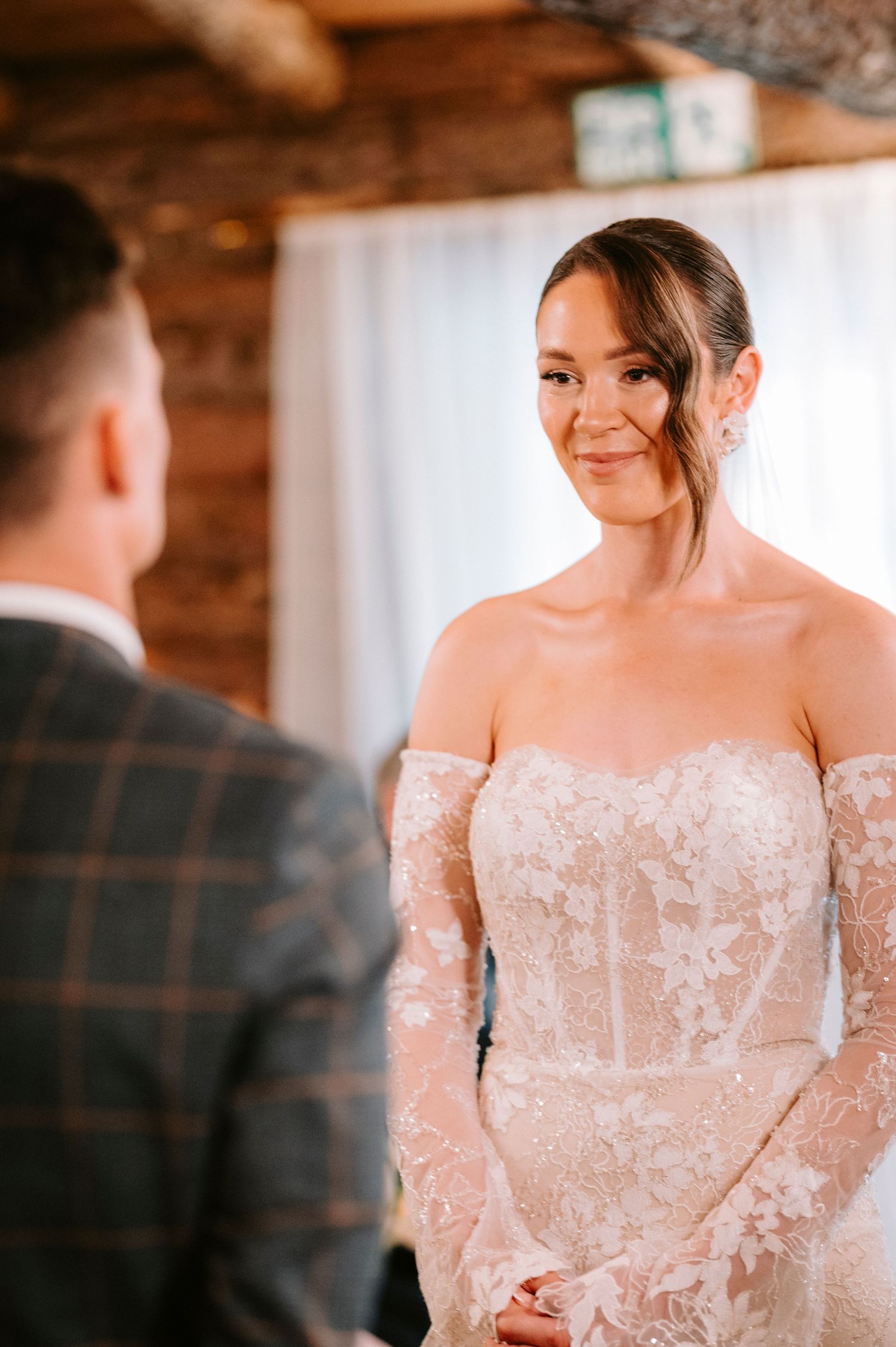 A bride in an off-shoulder lace wedding gown stands in front of a rustic wooden wall.