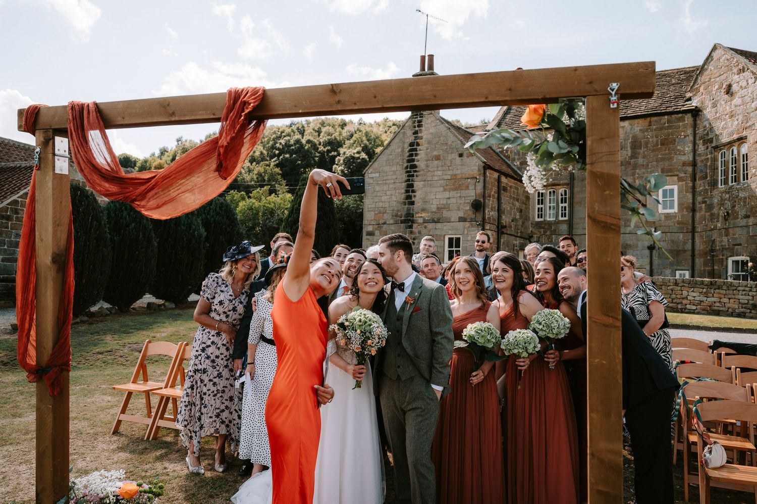 Wedding guests gathered under wooden pergola decorated with red fabric and floral arrangements.