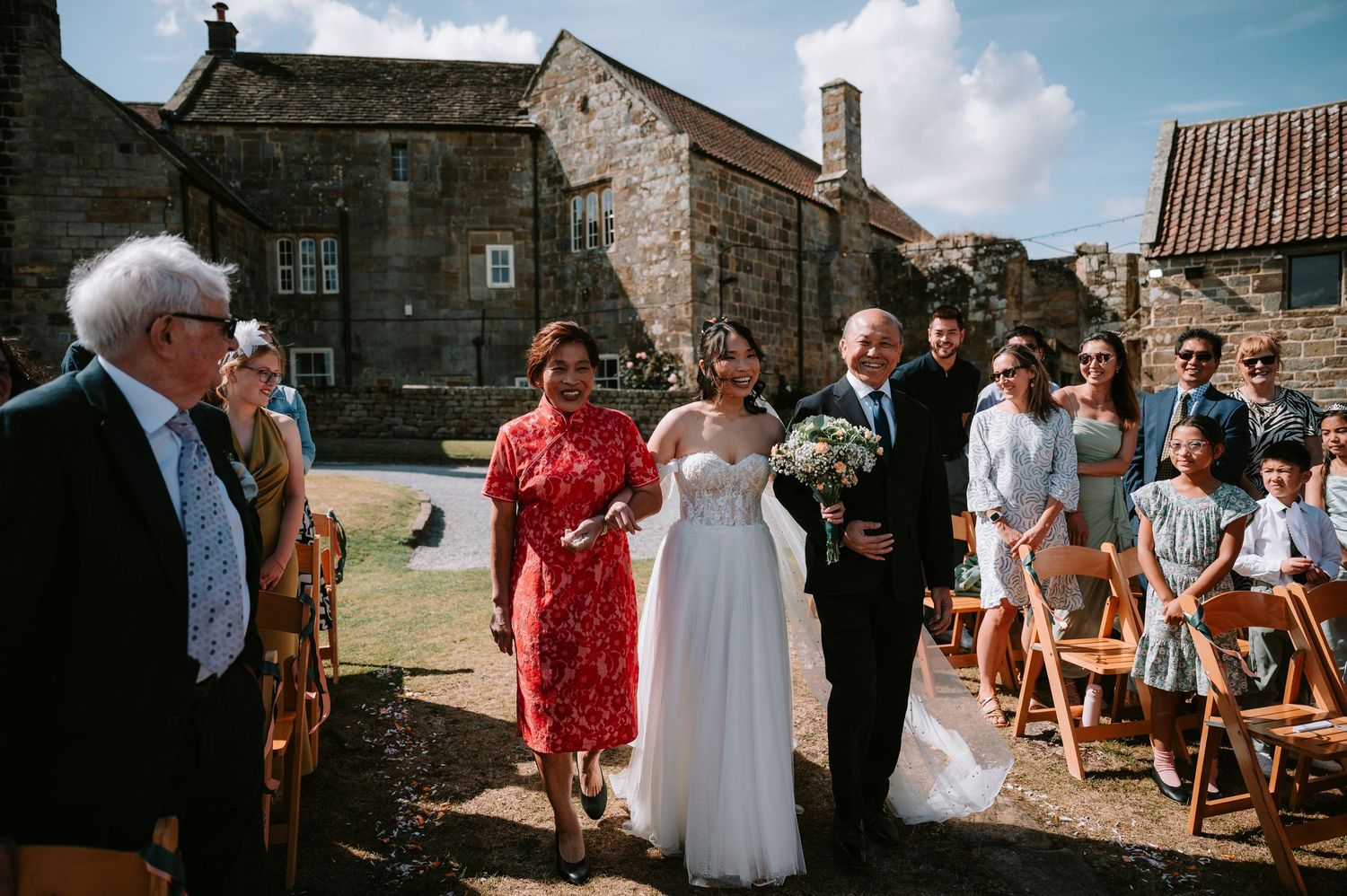 Outdoor wedding ceremony with guests seated on wooden chairs at a rustic stone venue.