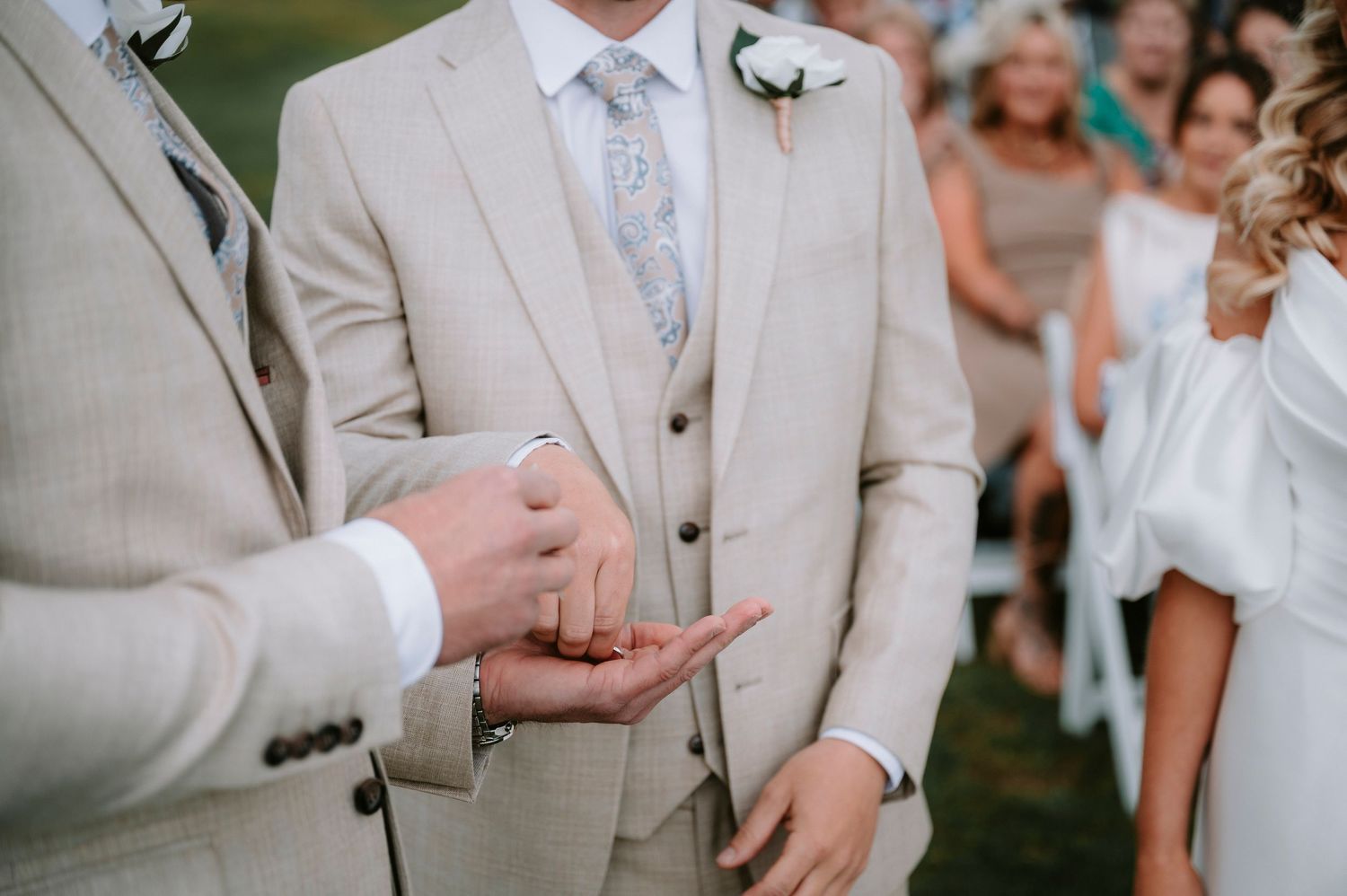 Groomsmen in light beige suits and blue ties stand together during an elegant outdoor wedding ceremony.