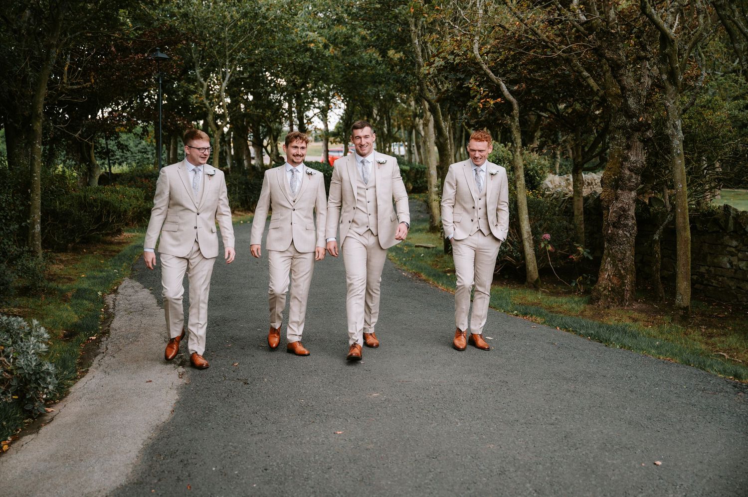 Groomsmen walking in formation down a tree-lined path in matching light-colored suits.