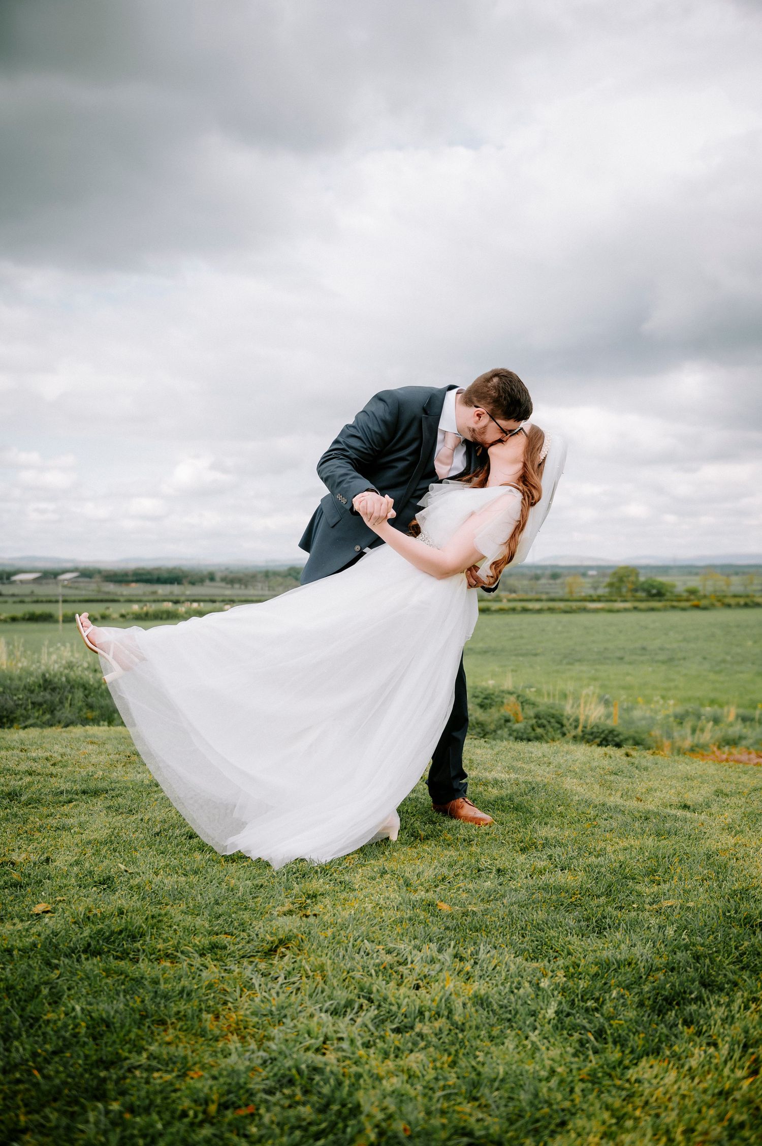 Romantic dip kiss on a scenic hilltop with rolling green countryside in the background.