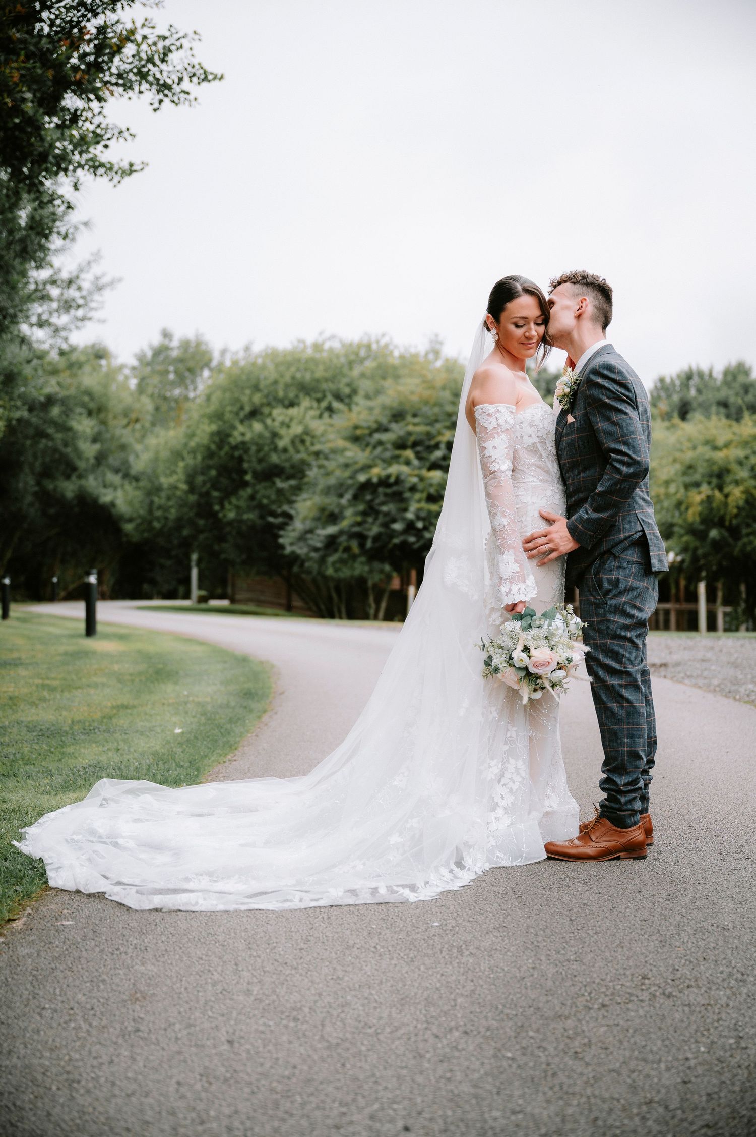 Wedding couple embraces on garden path with bride holding delicate bouquet and wearing flowing lace gown.