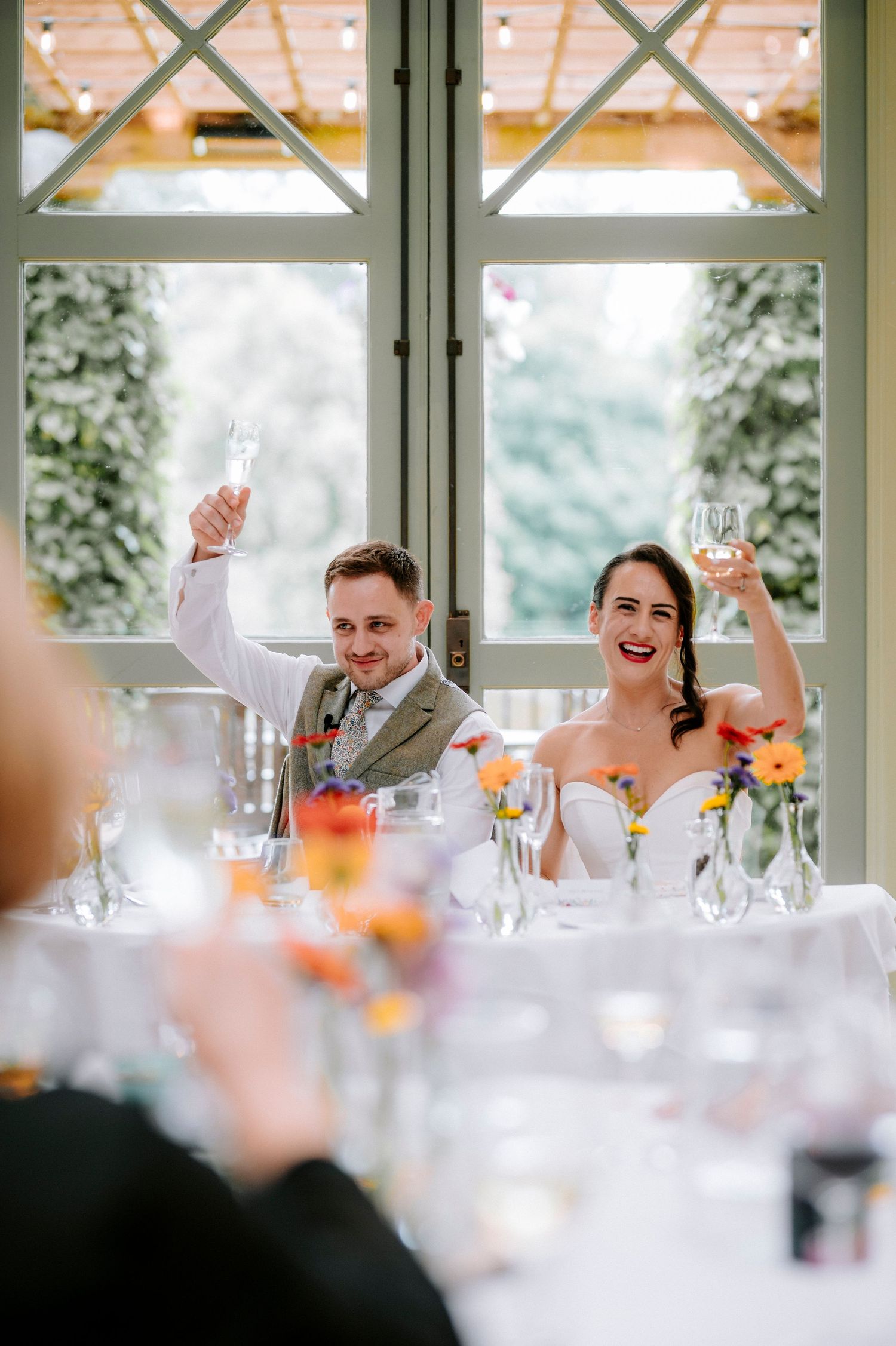 Wedding guests laugh and celebrate during reception toasts in sunlit venue.