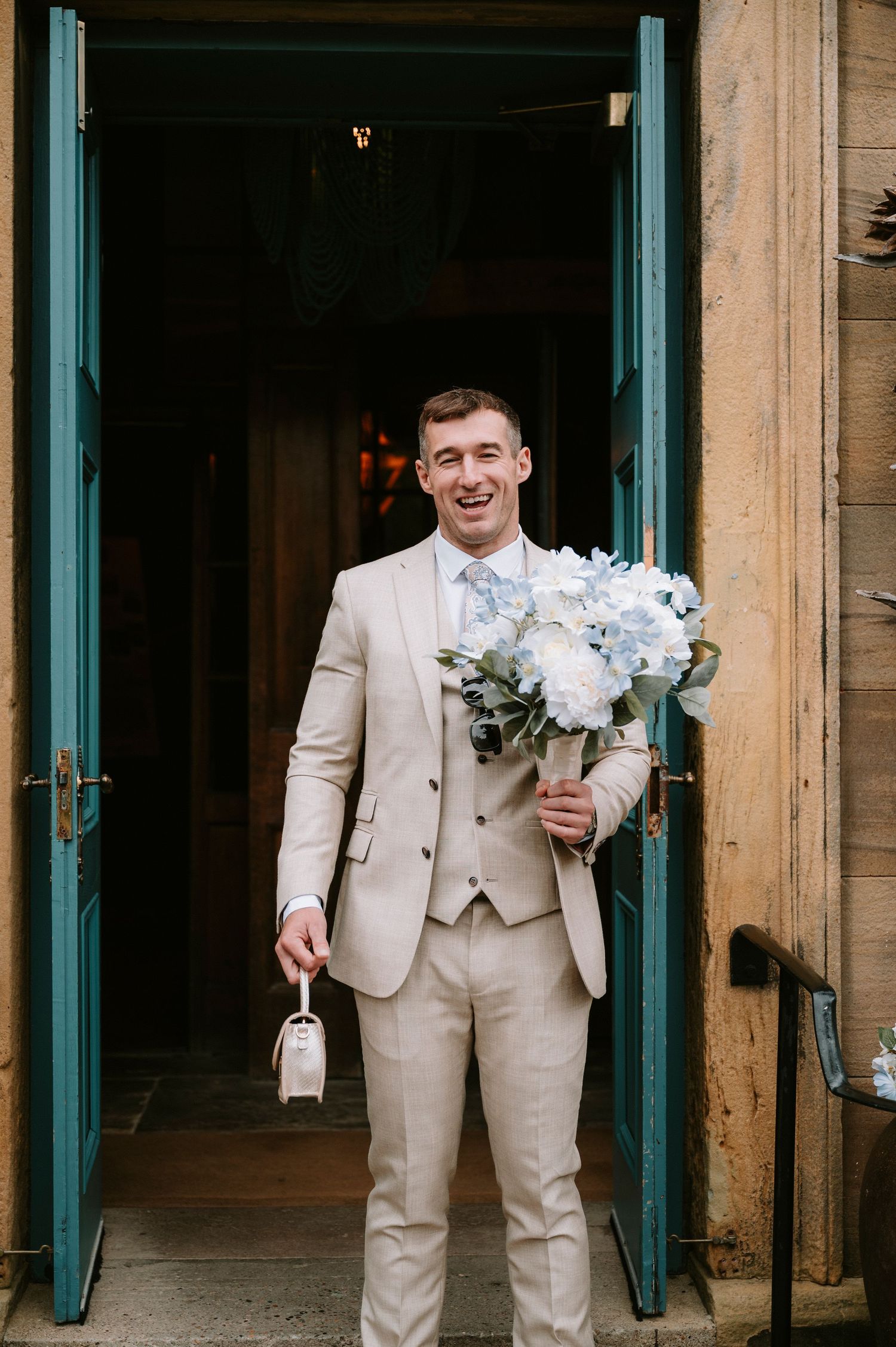 Groomsmen in beige suits holding white floral bouquets stand in front of green doors.