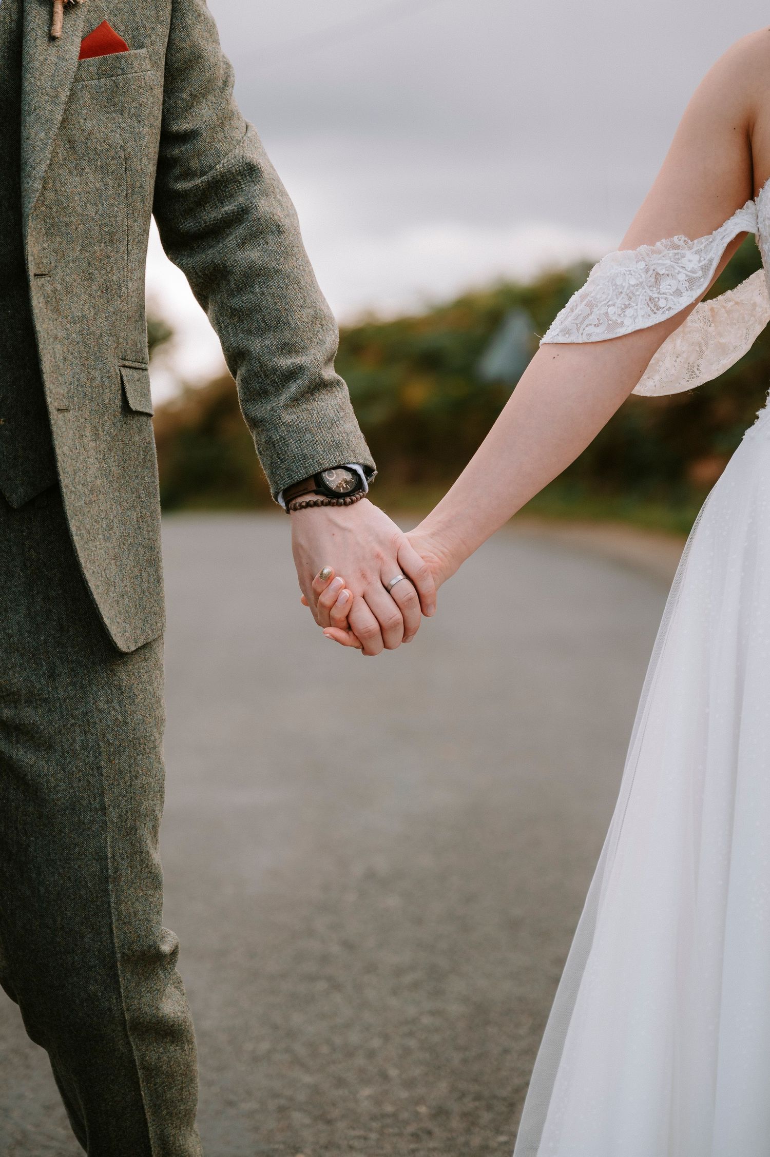 A couple in wedding attire holds hands while standing on a paved path, with the groom wearing a grey suit.
