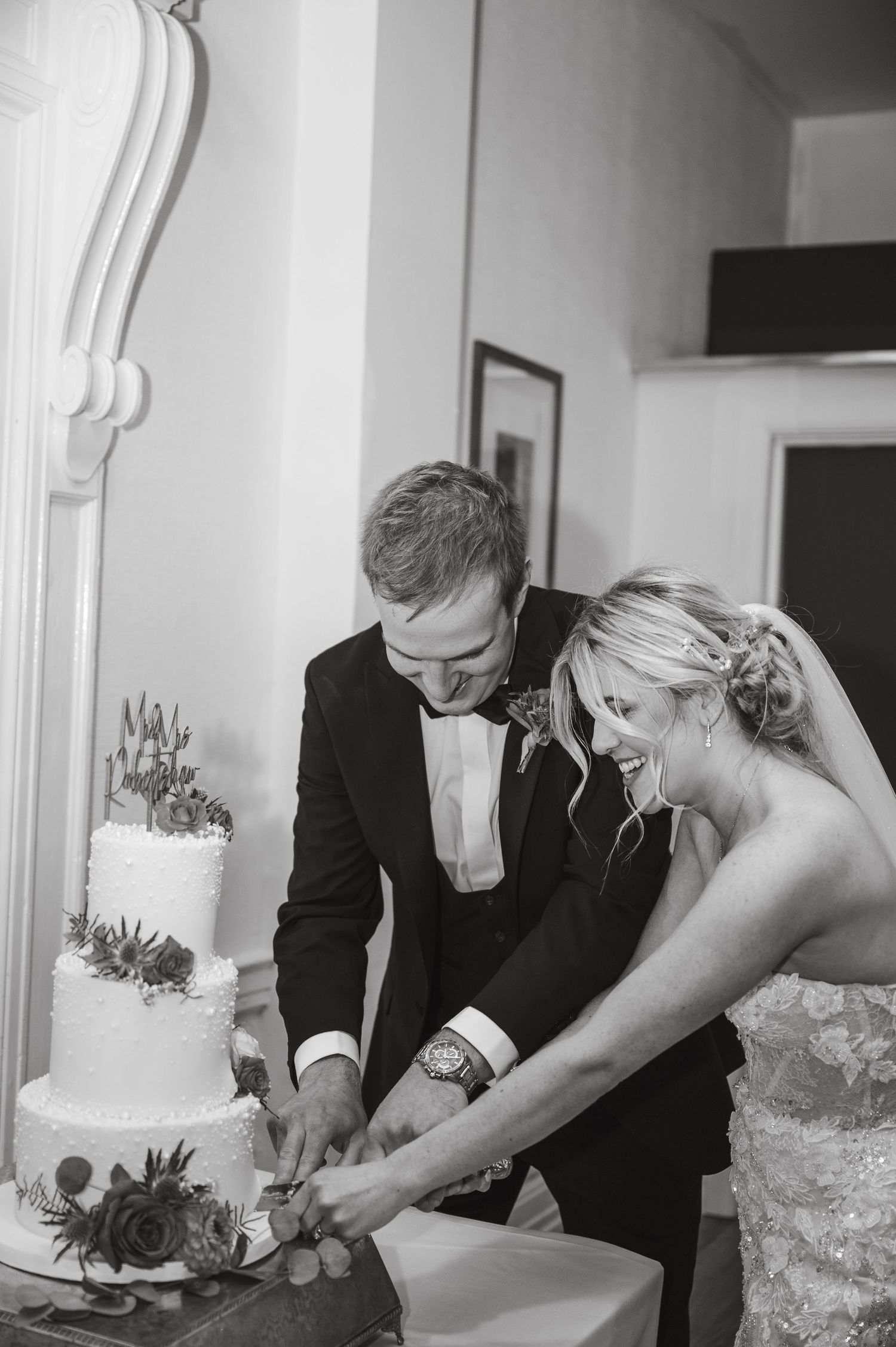 A couple works together to cut their tiered white wedding cake at their reception.