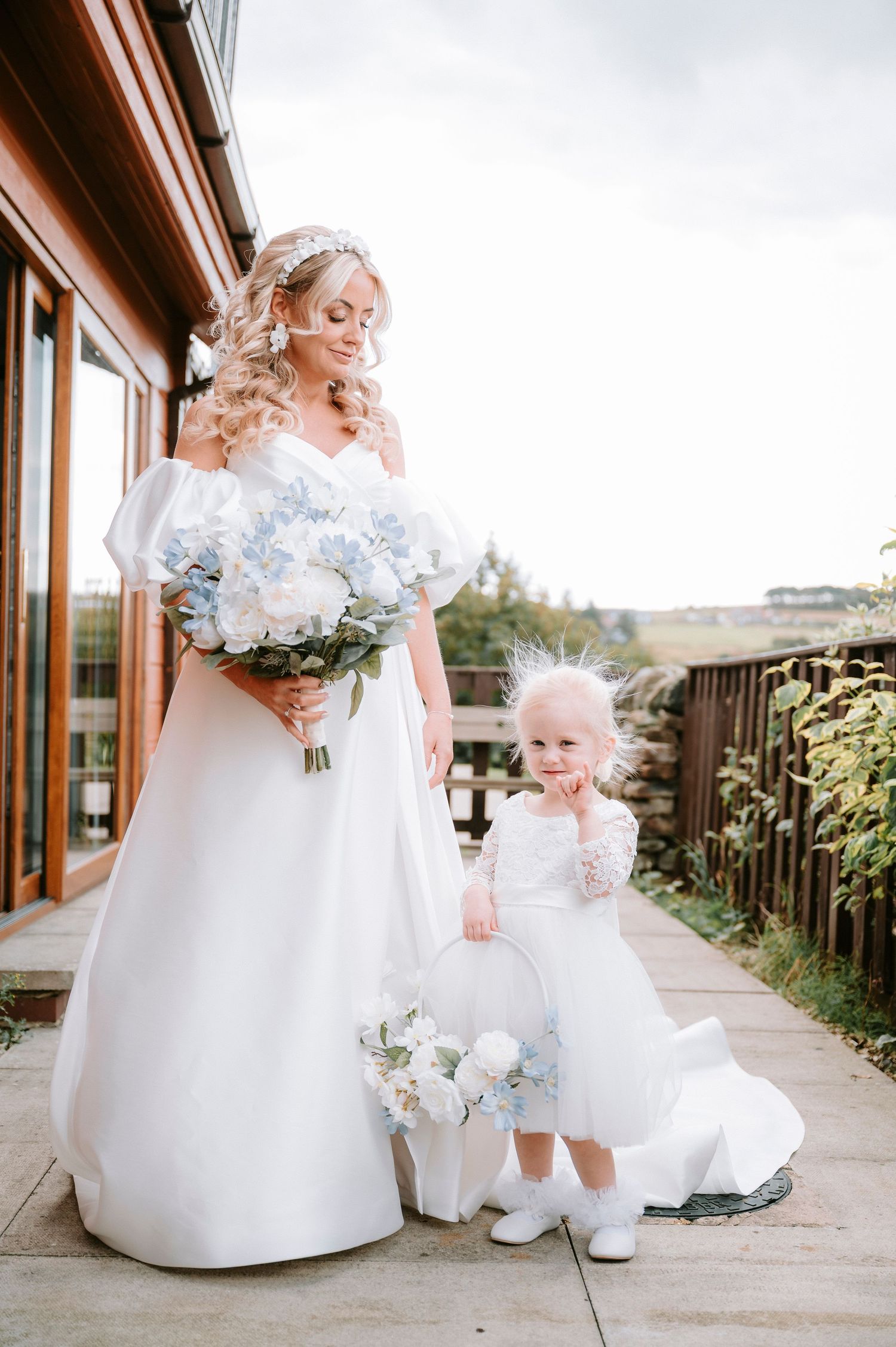 Bride in white gown stands with young flower girl on outdoor terrace holding matching blue bouquets.