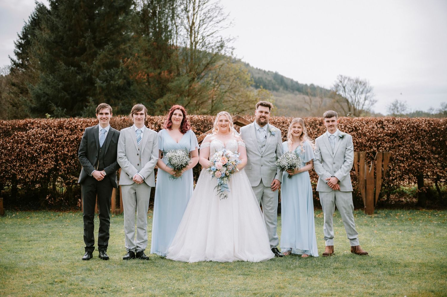 A wedding party in light blue and grey attire poses together on a lawn with mountain views.