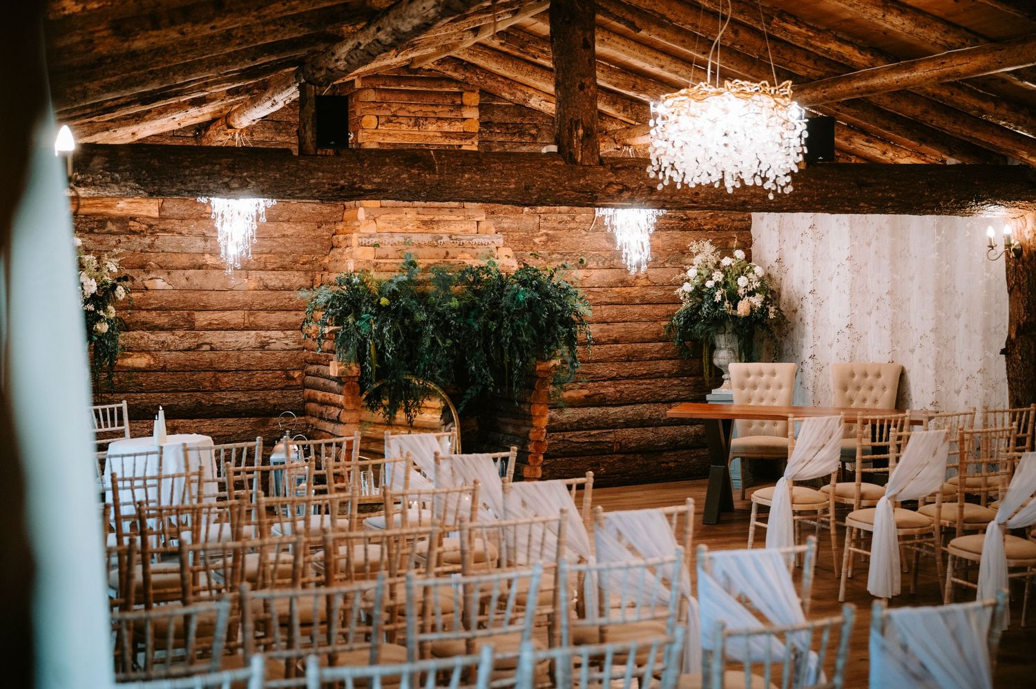 Empty rustic wedding ceremony venue with wooden chairs and chandeliers hanging from exposed beam ceiling.