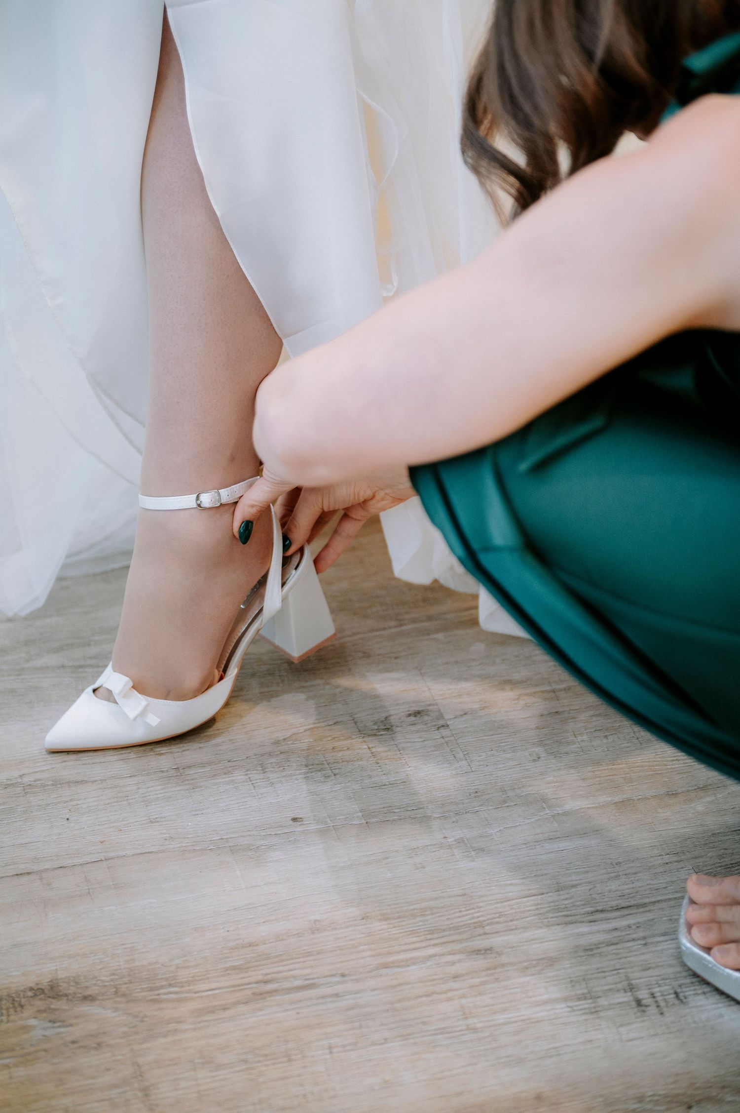 Someone helps the bride put on elegant white wedding shoes on wooden flooring.