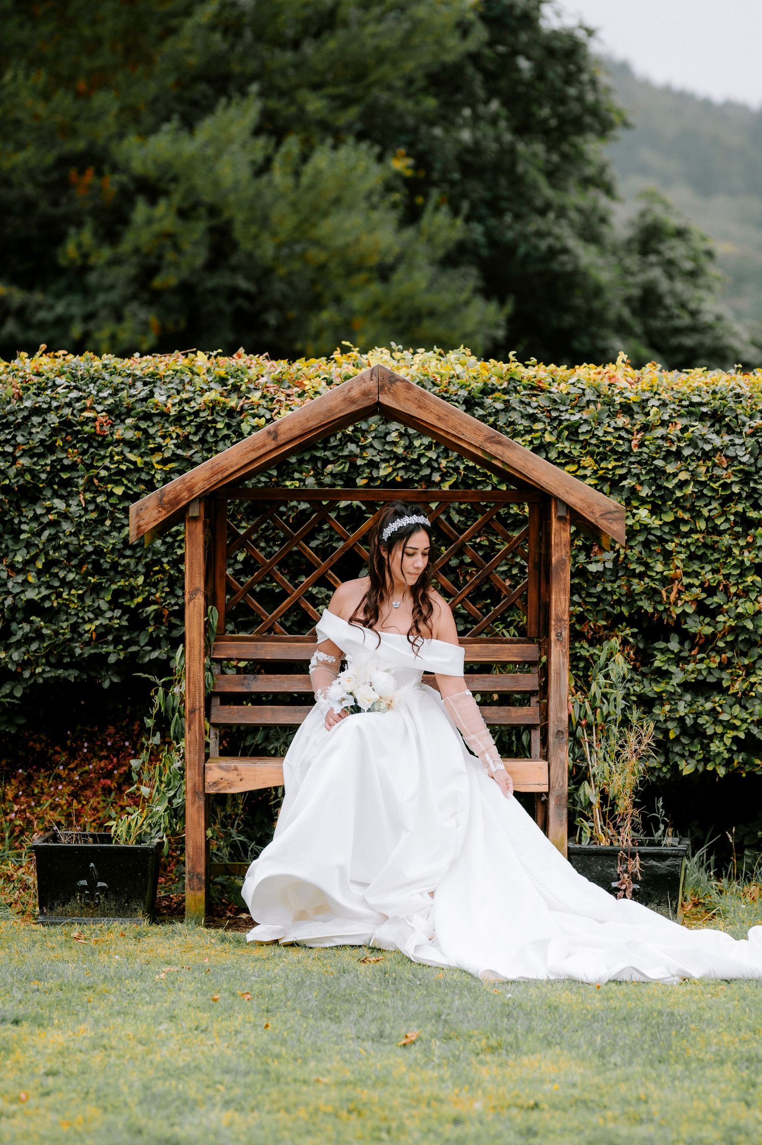 A bride in a white gown rests on a wooden garden bench surrounded by green hedges.