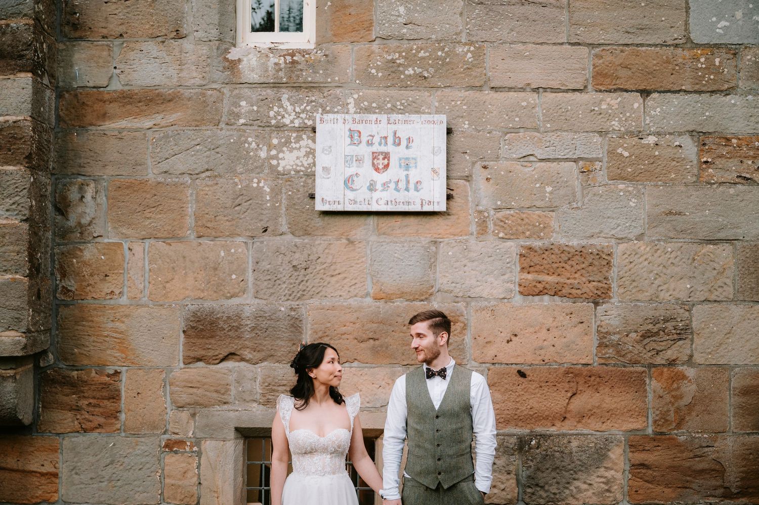 A couple poses in formal wedding attire against a historic stone wall backdrop.