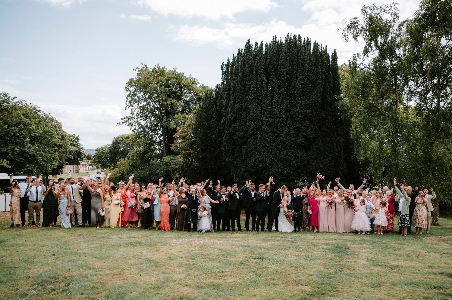 Wide shot of wedding guests assembled on grassy hillside for group photo.