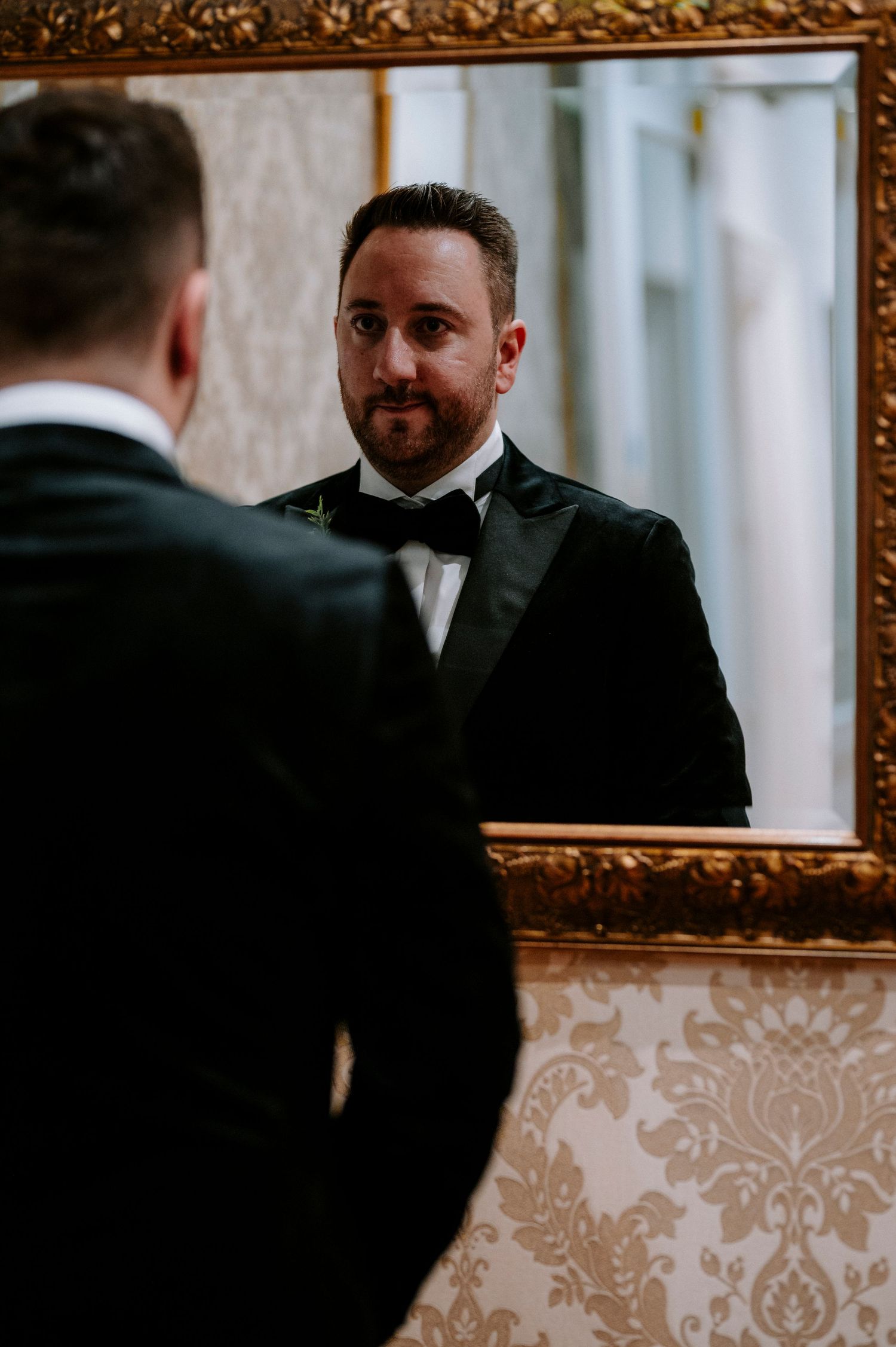 Dark suited figures reflected in an ornate gold-framed mirror against damask wallpaper.