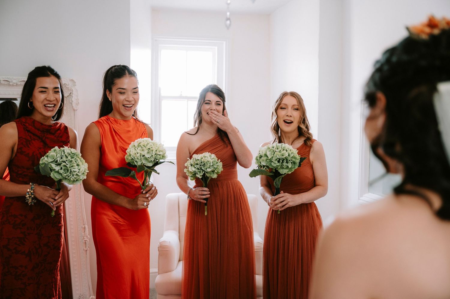 Bridesmaids in red and burgundy dresses sharing a joyful moment in a bright white room.