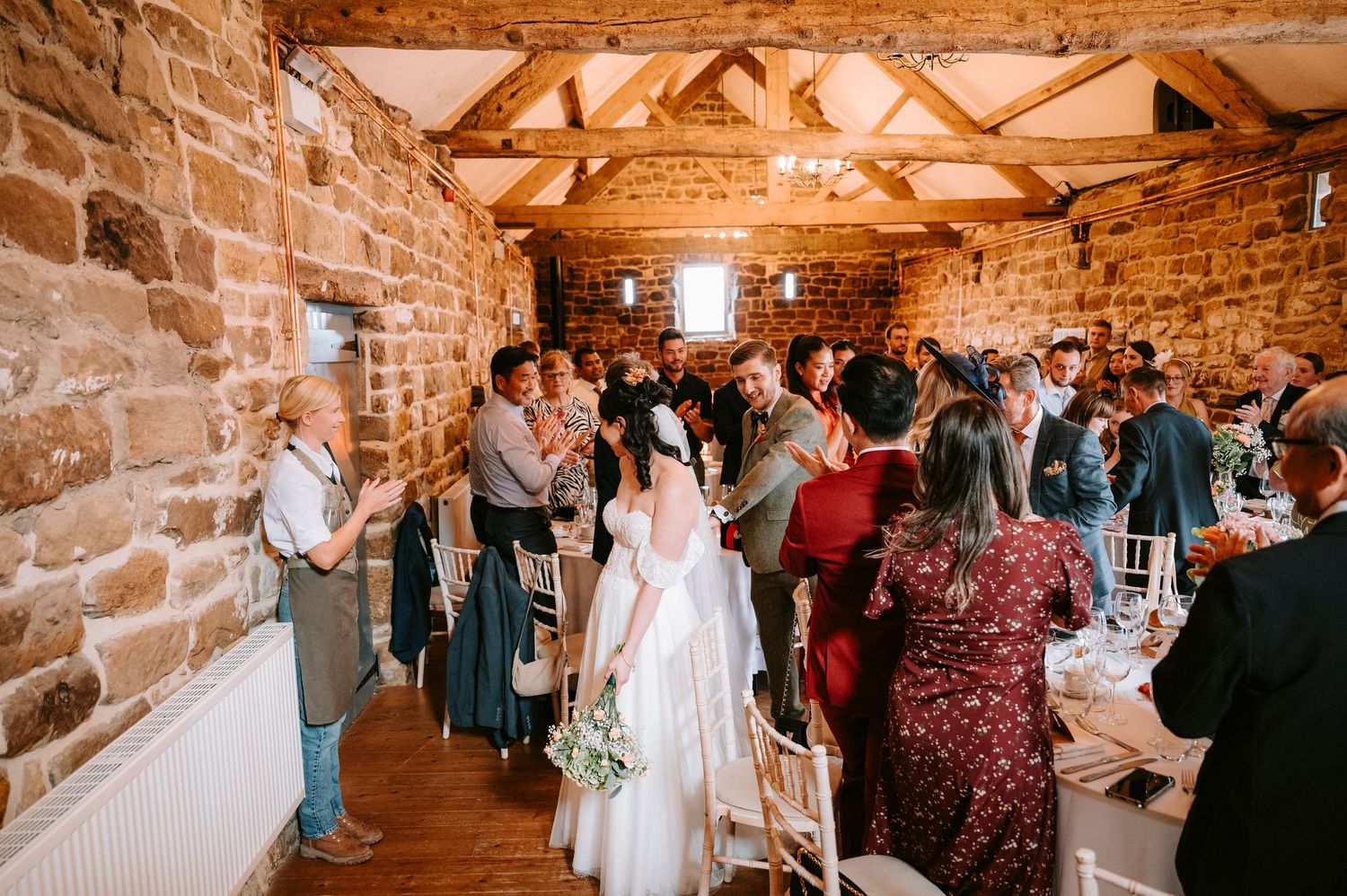 Wedding guests celebrate in a rustic stone-walled venue with exposed wooden beams and warm lighting.