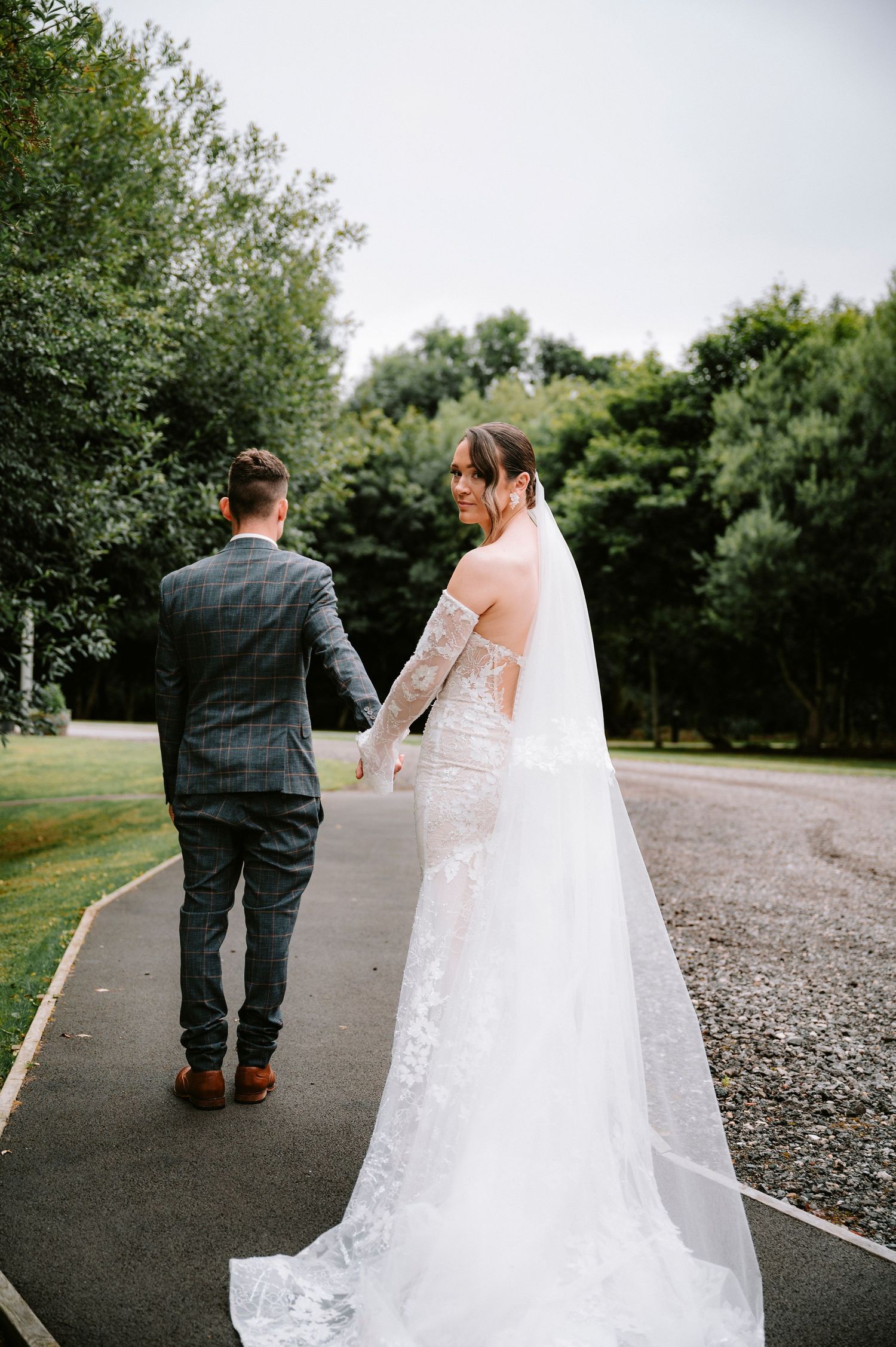 Bride in lace gown with flowing veil walks with groom along outdoor path surrounded by greenery.