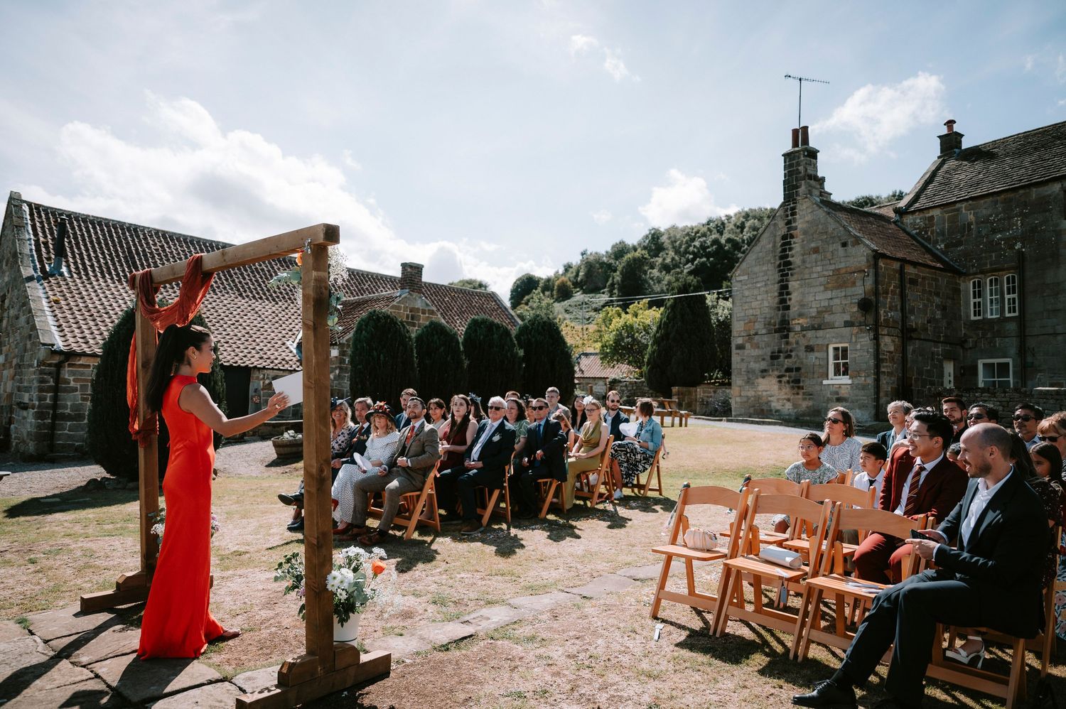 Outdoor wedding ceremony with guests seated on wooden chairs facing a rustic arch and stone buildings.