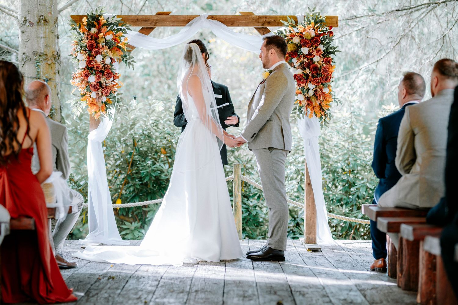 Wedding ceremony under a floral arch with autumn-colored decorations and wooden benches.