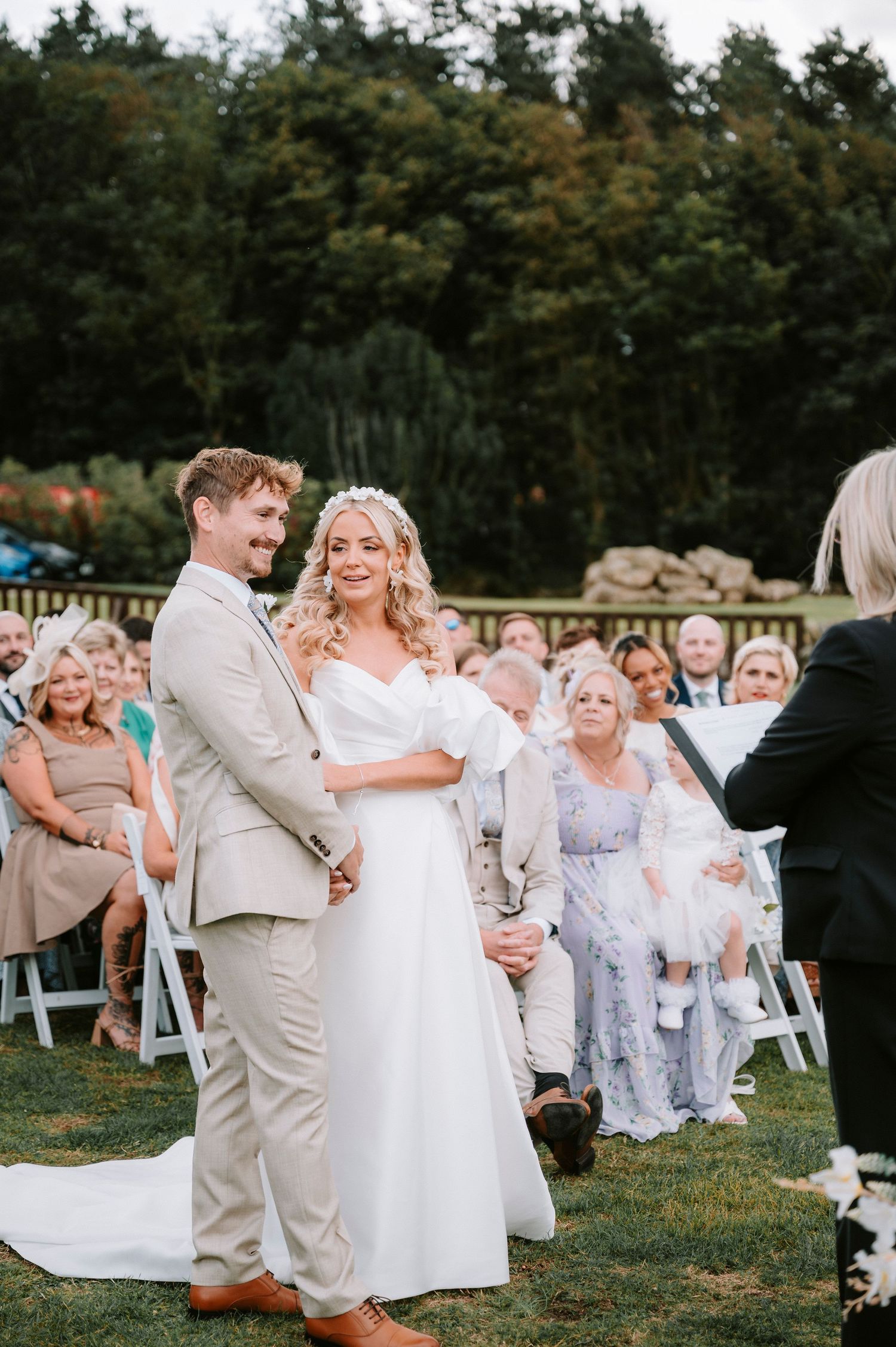 Outdoor wedding ceremony with guests seated on white chairs on a grassy lawn.