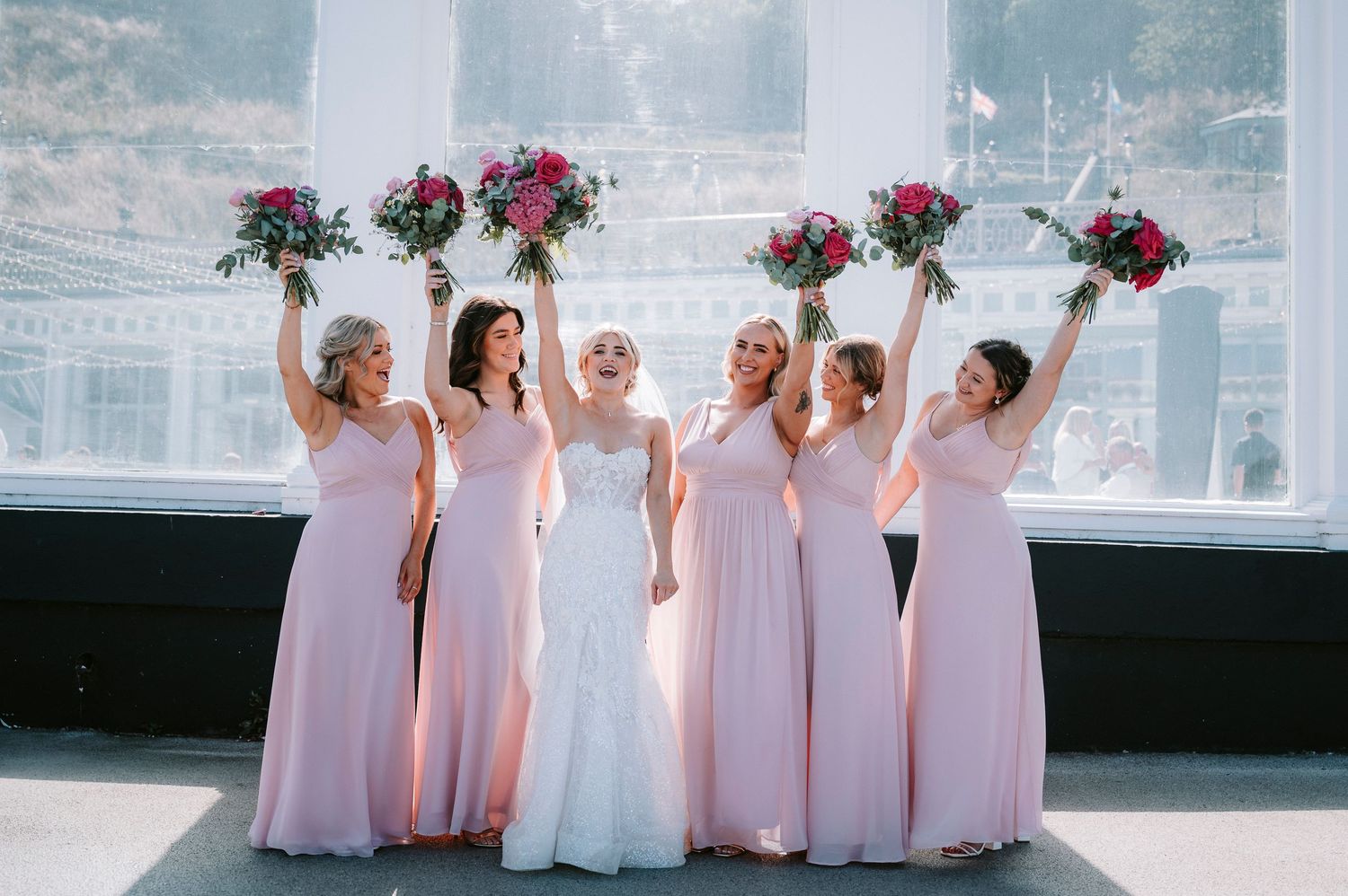 Bridesmaids in pink dresses holding bouquets pose together against a bright backdrop.