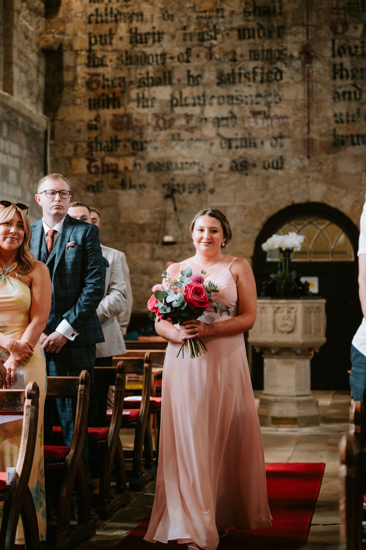 Bridesmaids in pink dresses stand in historic church with ancient text on stone walls.