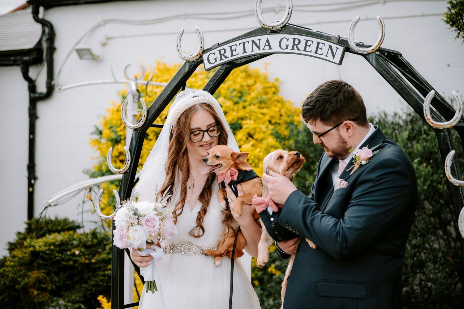Wedding ceremony under a rustic Gretna Green archway with yellow flowers blooming in background.
