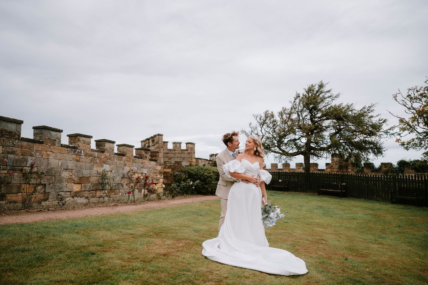 Wedding couple poses along stone castle wall pathway during outdoor photo session.