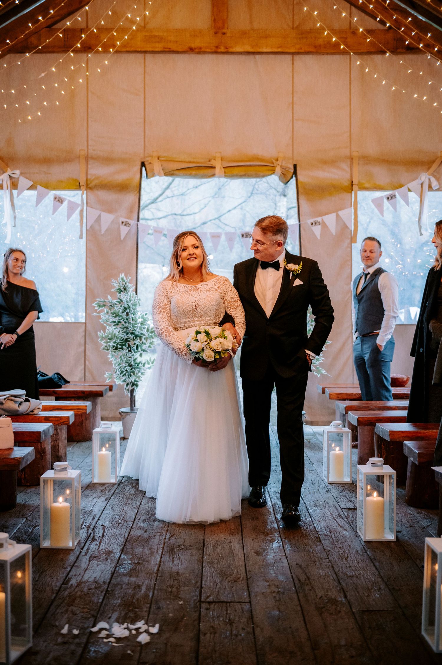 A couple walks down the aisle in a rustic barn venue decorated with string lights and candles.