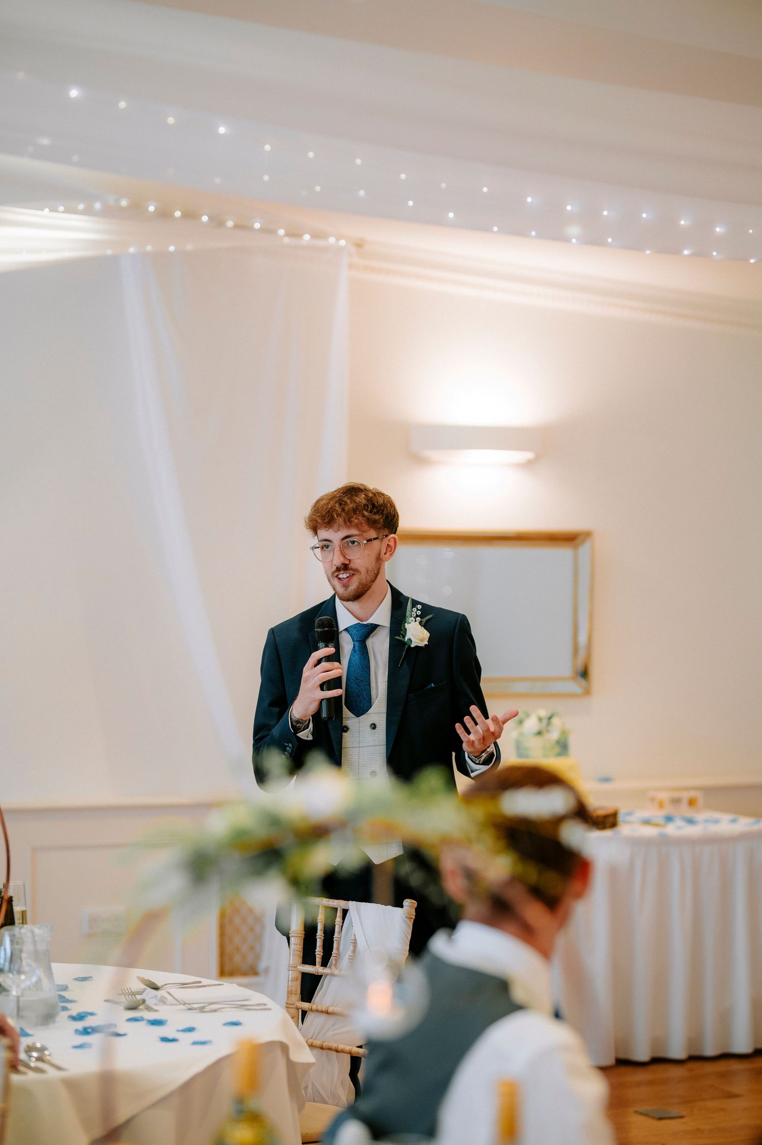 Wedding reception guests laughing and enjoying speeches in an elegant white room with fairy lights.