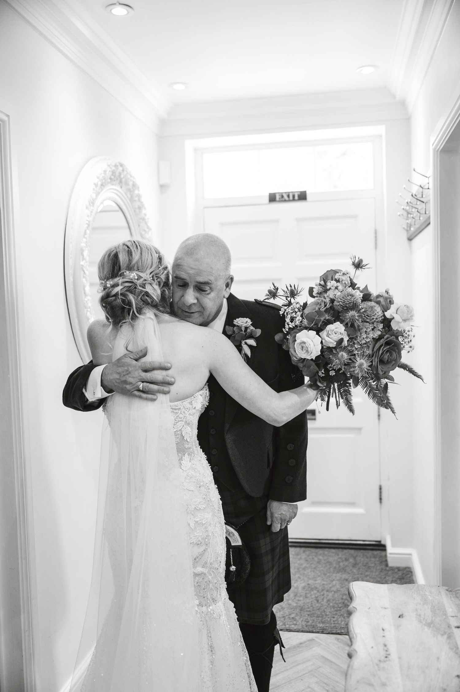 Emotional first look moment in a bright white hallway with floral bouquet.