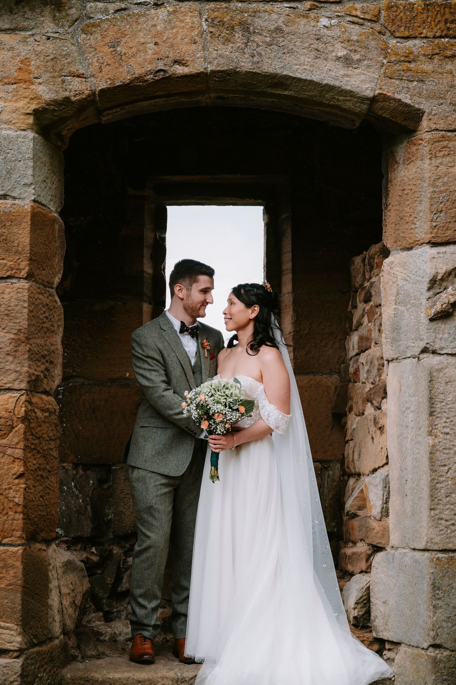 A romantic wedding photoshoot in stone archways with a couple in a white dress and grey suit.