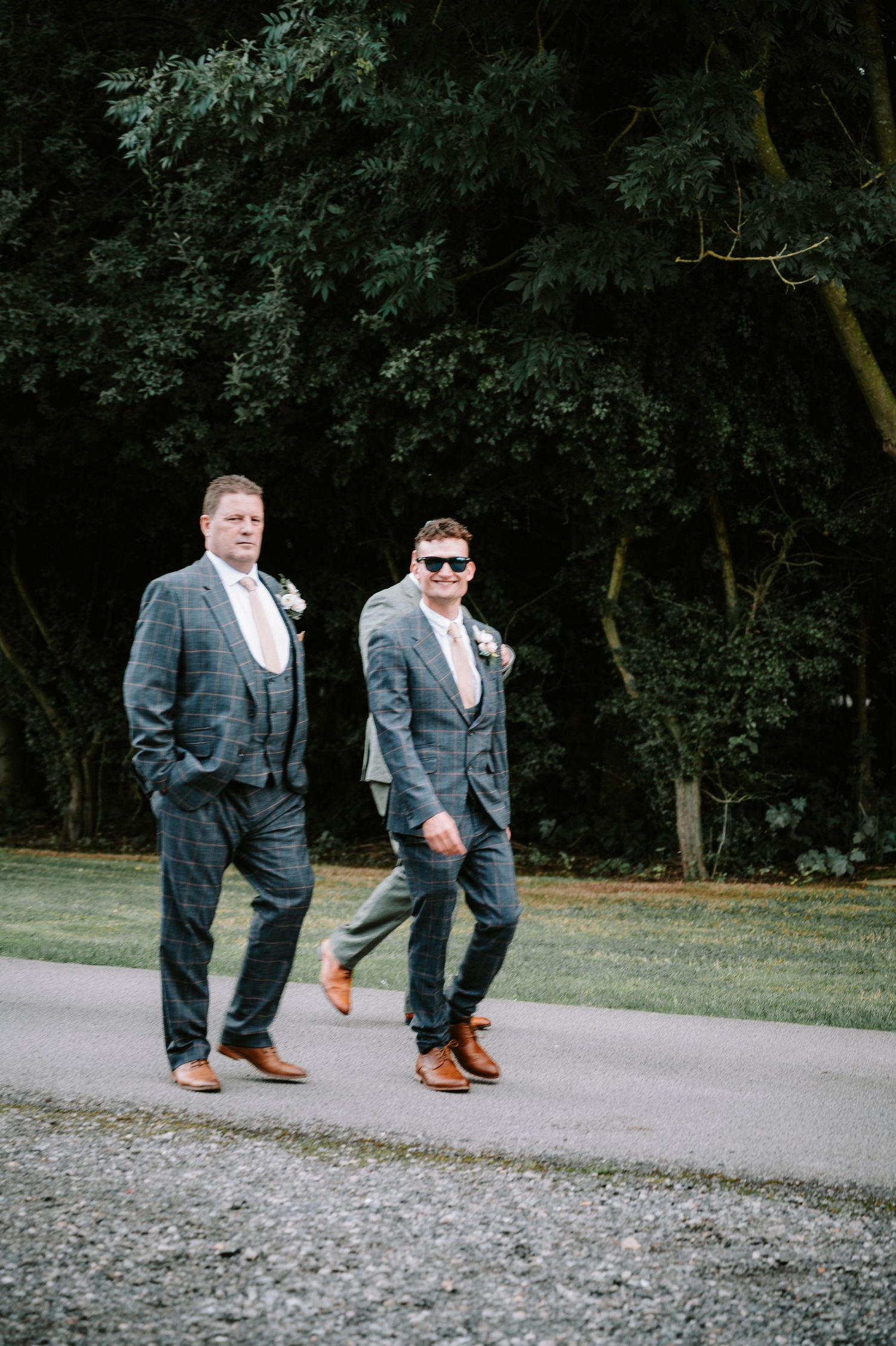 Groomsmen in grey suits and brown shoes walking on gravel path.