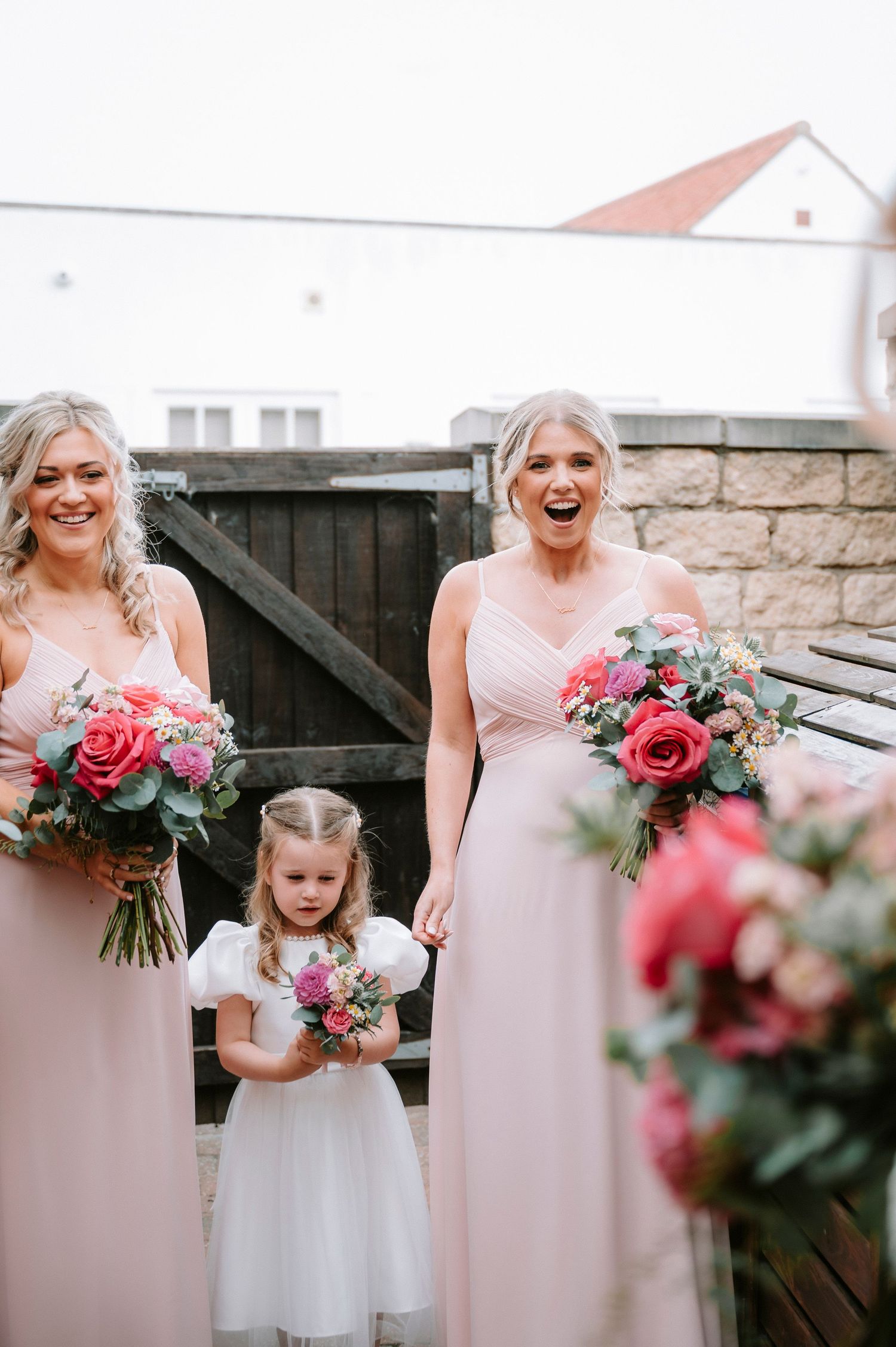 Bridesmaids in blush pink dresses holding vibrant rose bouquets against stone wall.
