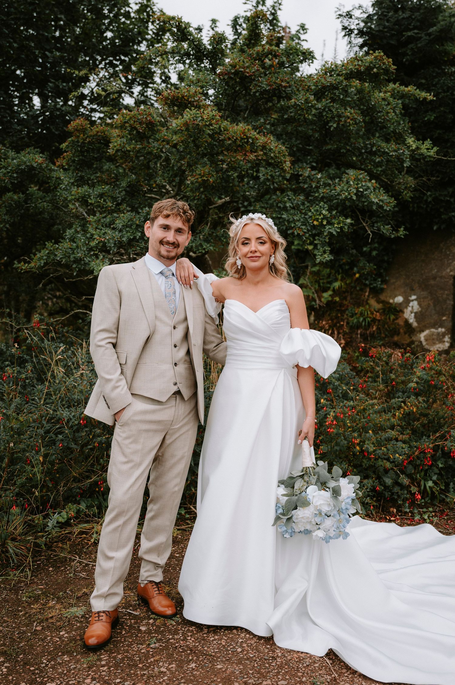 Wedding couple poses in garden setting with bride in white gown and groom in beige suit.