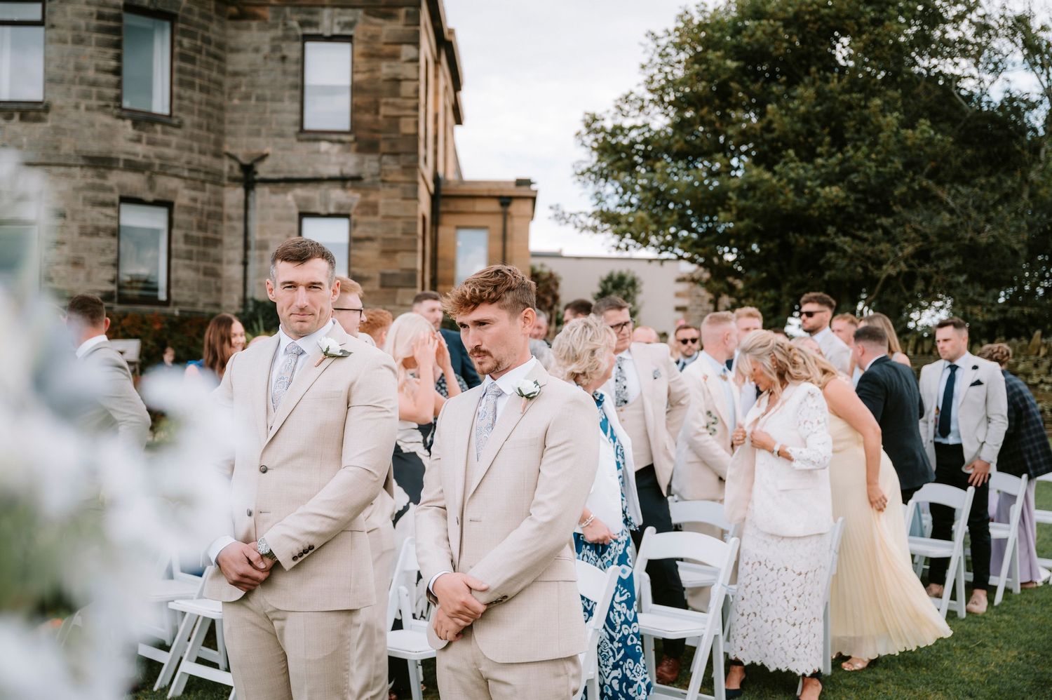 Wedding guests seated in white chairs watch an outdoor ceremony with groomsmen in beige suits.
