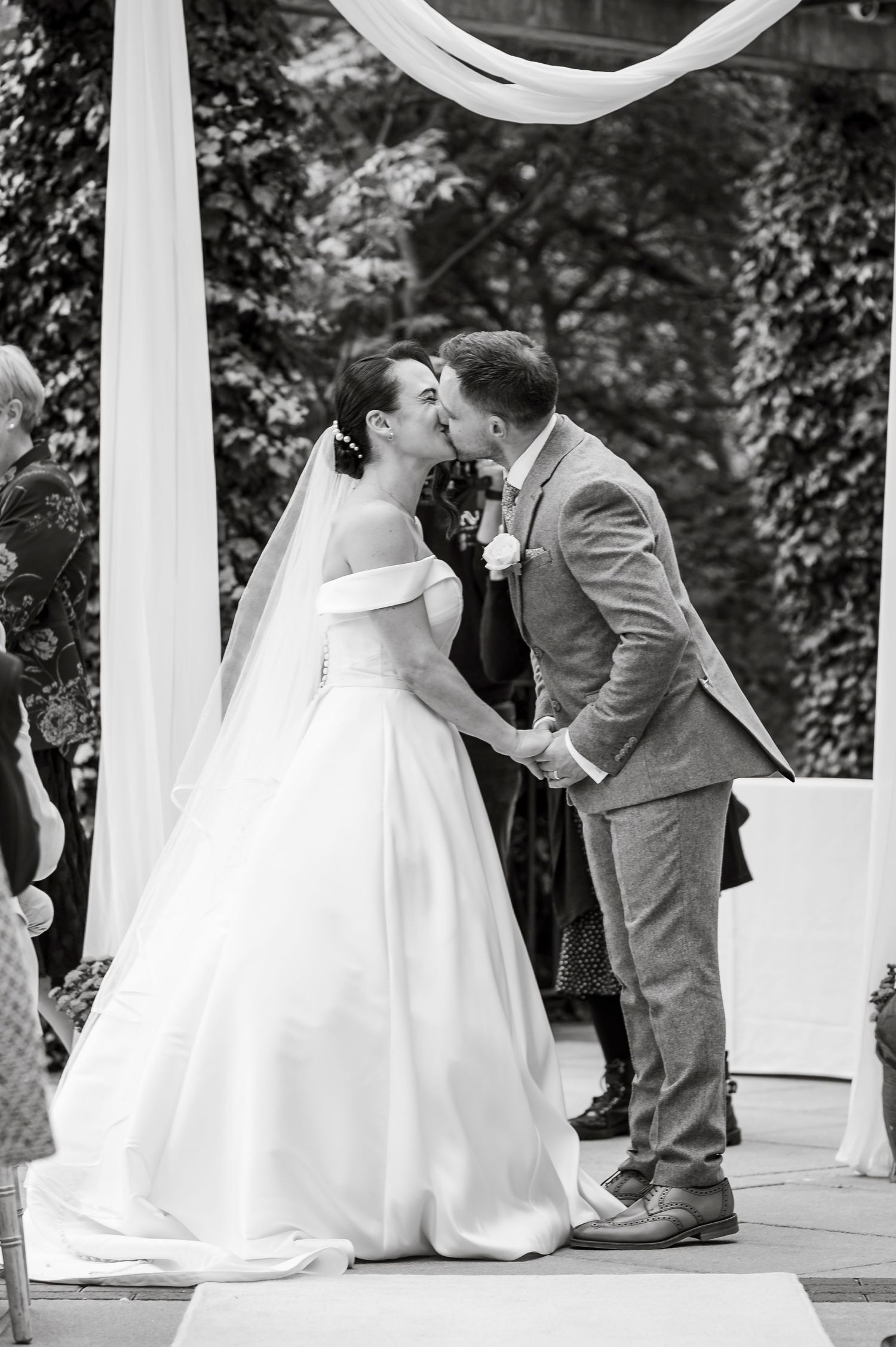 Wedding couple shares romantic first kiss under white draped ceremony arch.