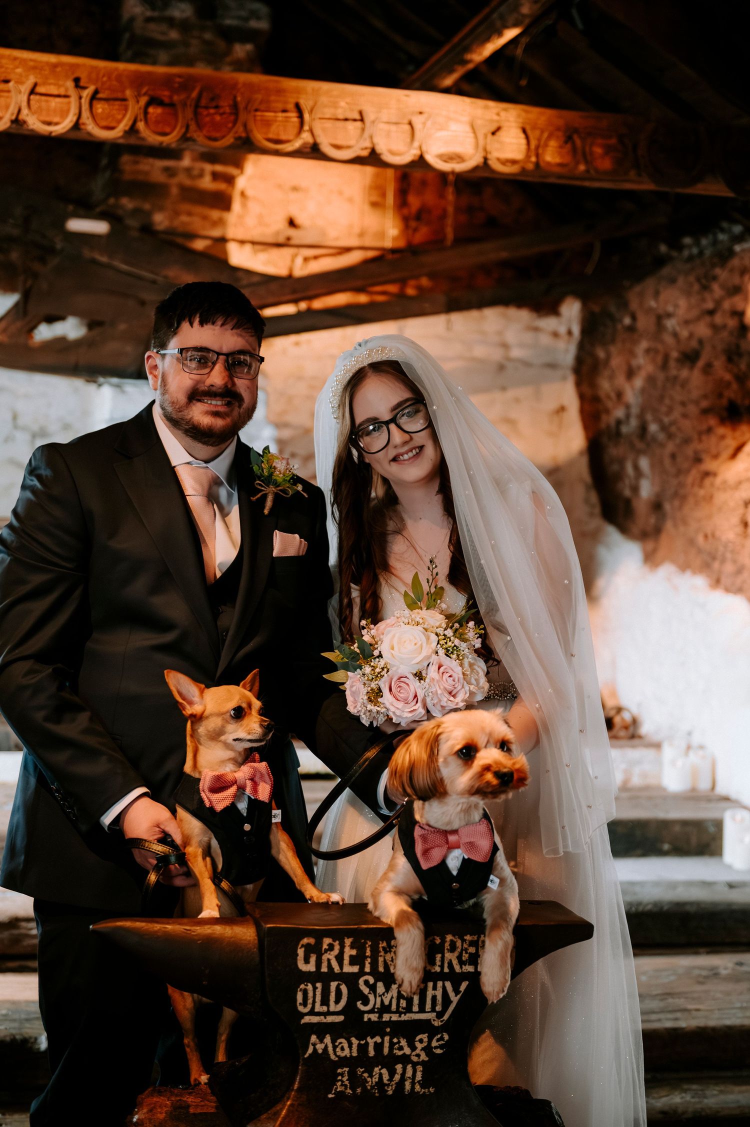 A wedding couple posing with their two small dogs at the Gretna Green Old Smithy anvil.